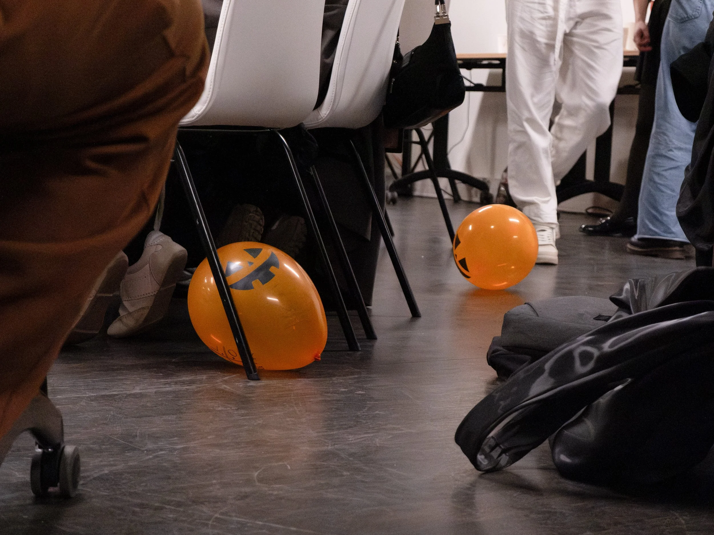 Des ballons d'Halloween orange avec des visages souriants et des détails de citrouille, posés sur le sol entre des chaises et des sacs, dans un espace intérieur fréquenté par plusieurs personnes.