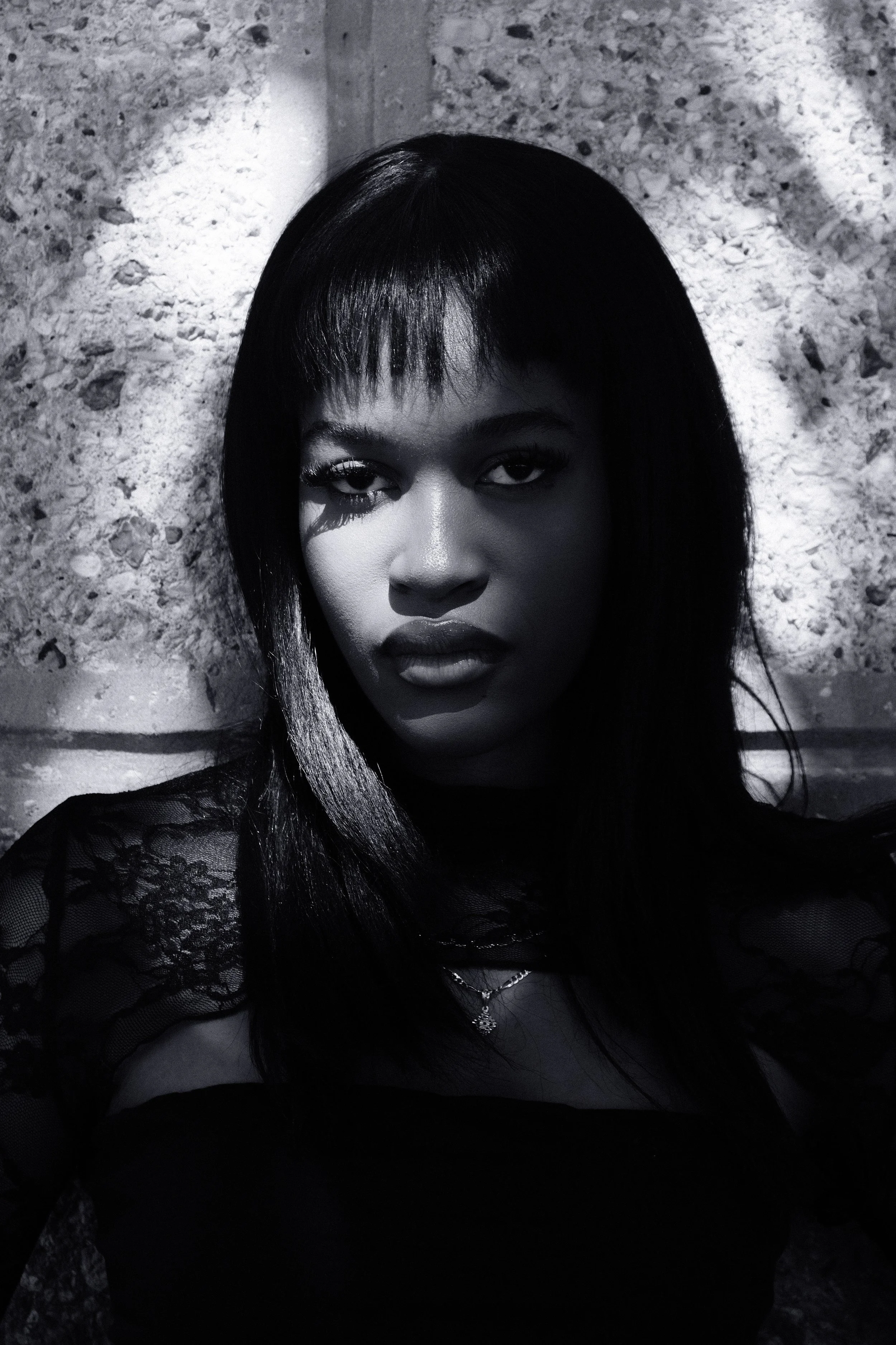 Portrait en noir et blanc d'une femme avec des cheveux longs et droits, portant un vêtement en dentelle et un collier, devant un mur en pierre.