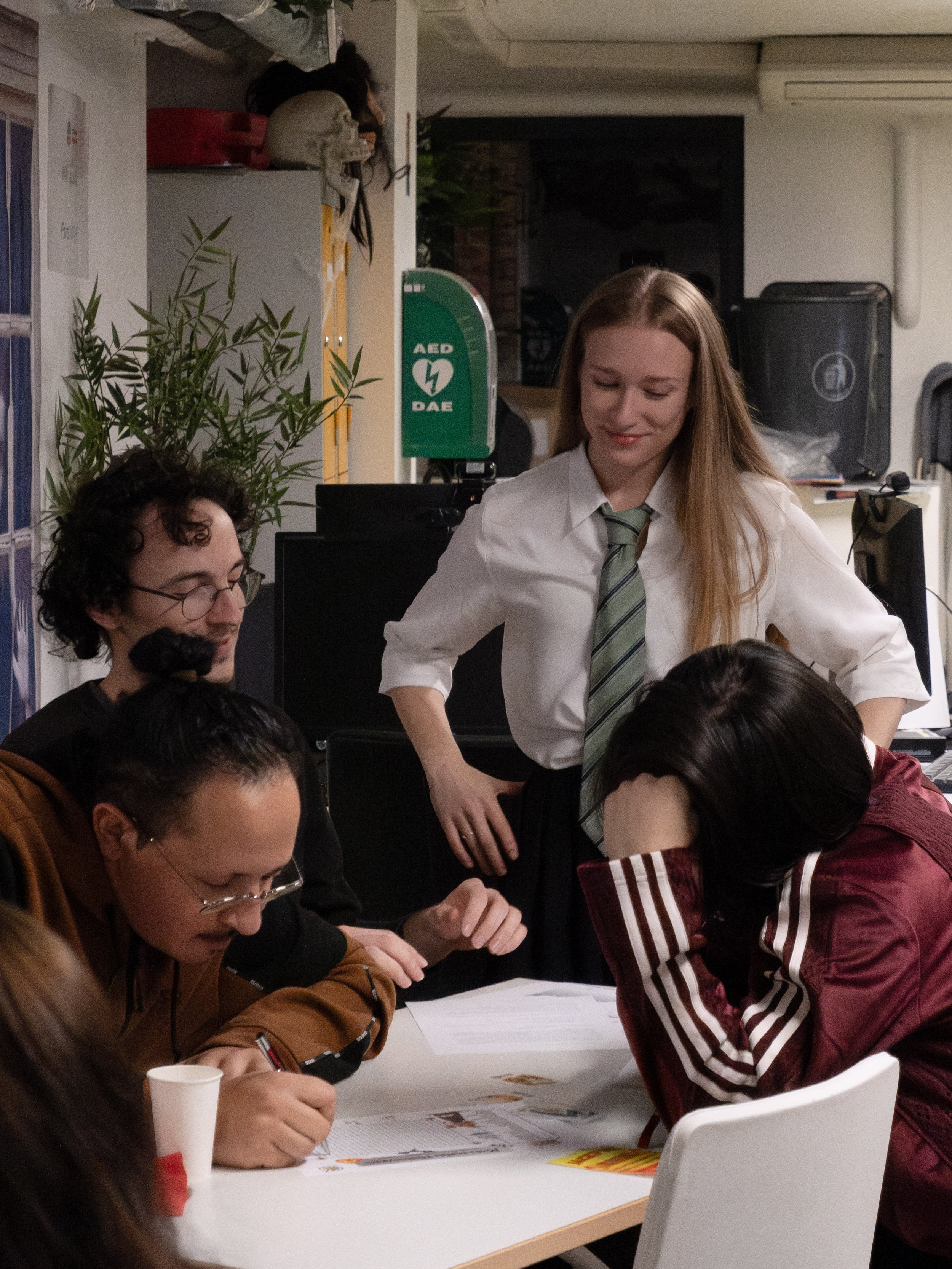 Groupes de jeunes assis autour d'une table, deux d'entre eux semblent concentrés sur des papiers ou des documents, tandis qu'une jeune femme se tient debout, regardant avec un sourire, dans un environnement intérieur avec une plante verte et du matér