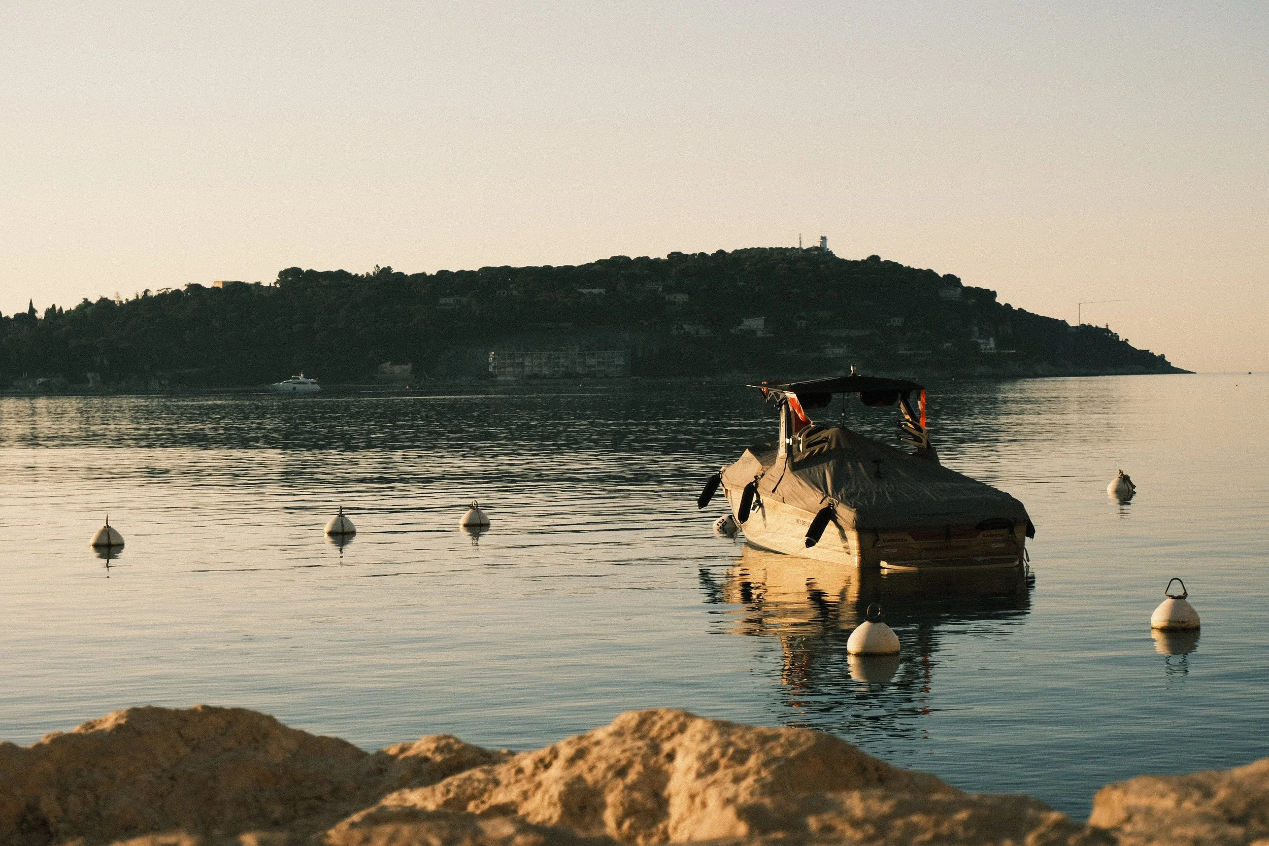 Bateau amarré dans une baie calme avec des bouées, vue sur une île ou une colline verdoyante au loin, au crépuscule.