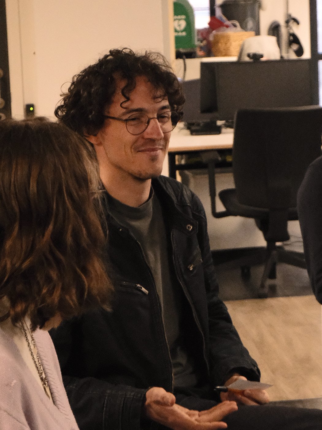 Un homme avec des cheveux bouclés noirs, portant des lunettes, souriant, assis dans un bureau ou espace de travail, avec une femme dont les cheveux bruns, vu de dos, à côté de lui.