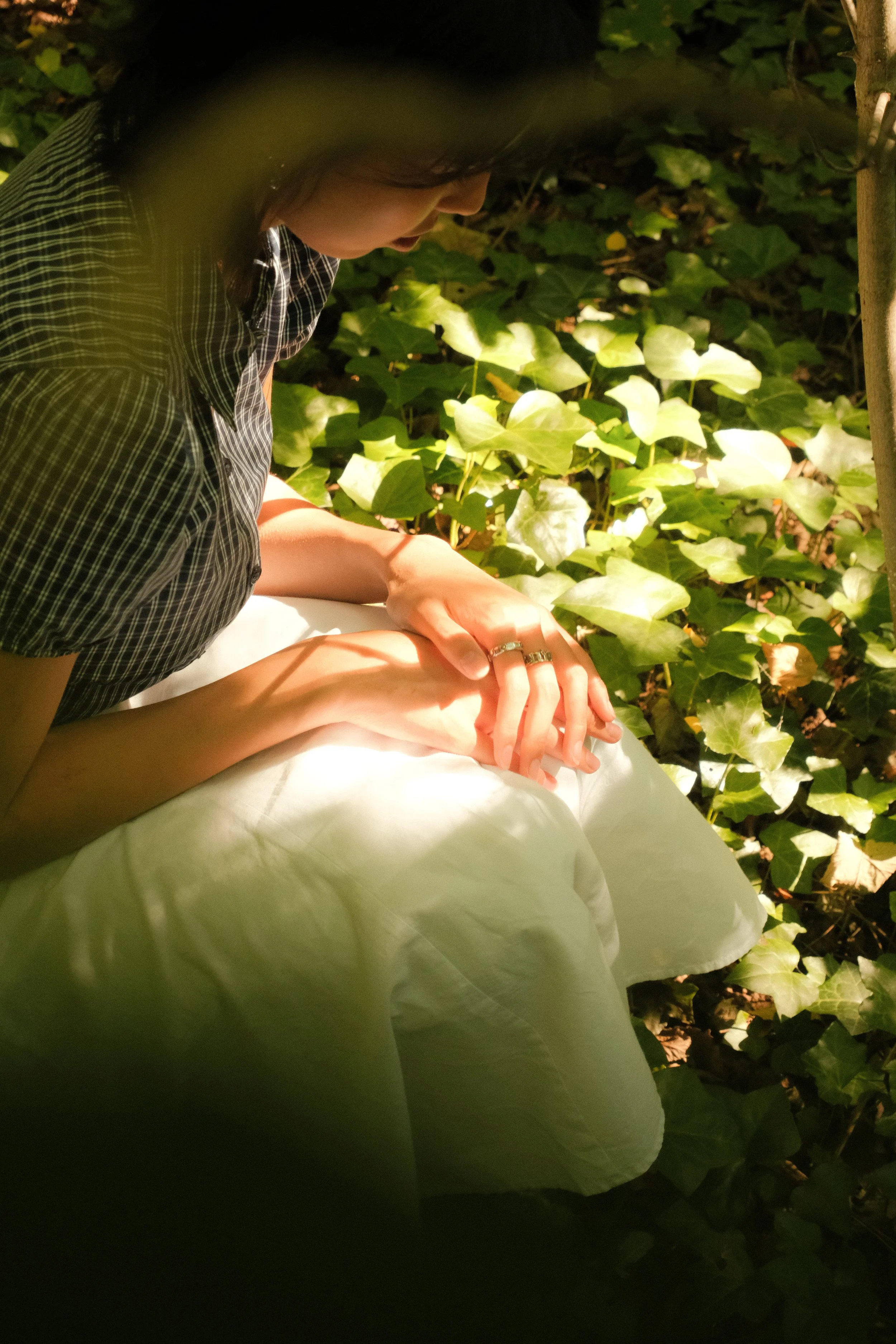 Femme assise dans un jardin, entourée de feuilles vertes, regardant vers le bas.