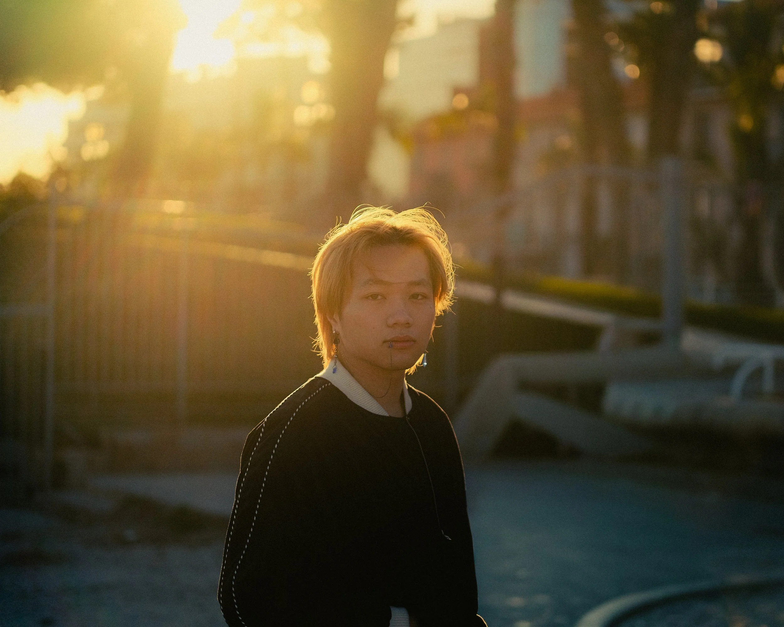 Un jeune homme avec des cheveux blonds ébouriffés, portant un sweat noir avec bordure blanche, regarde vers l'objectif dans une scène en plein air au coucher du soleil, avec des bâtiments et des arbres en arrière-plan.