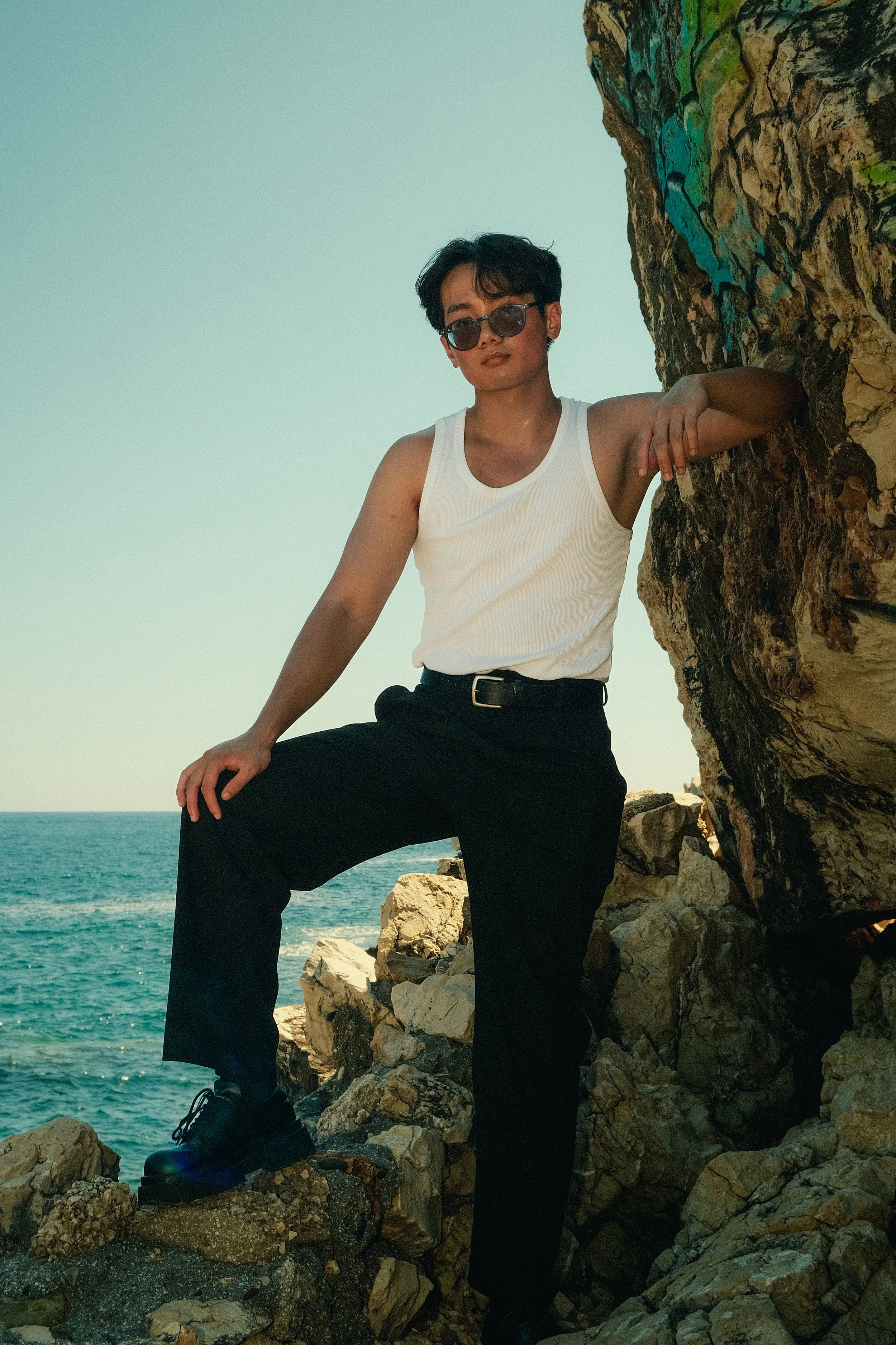Un jeune homme debout sur des rochers près de la mer, portant un t-shirt blanc, des pantalons noirs, des lunettes de soleil, avec la mer en arrière-plan et un ciel clair.