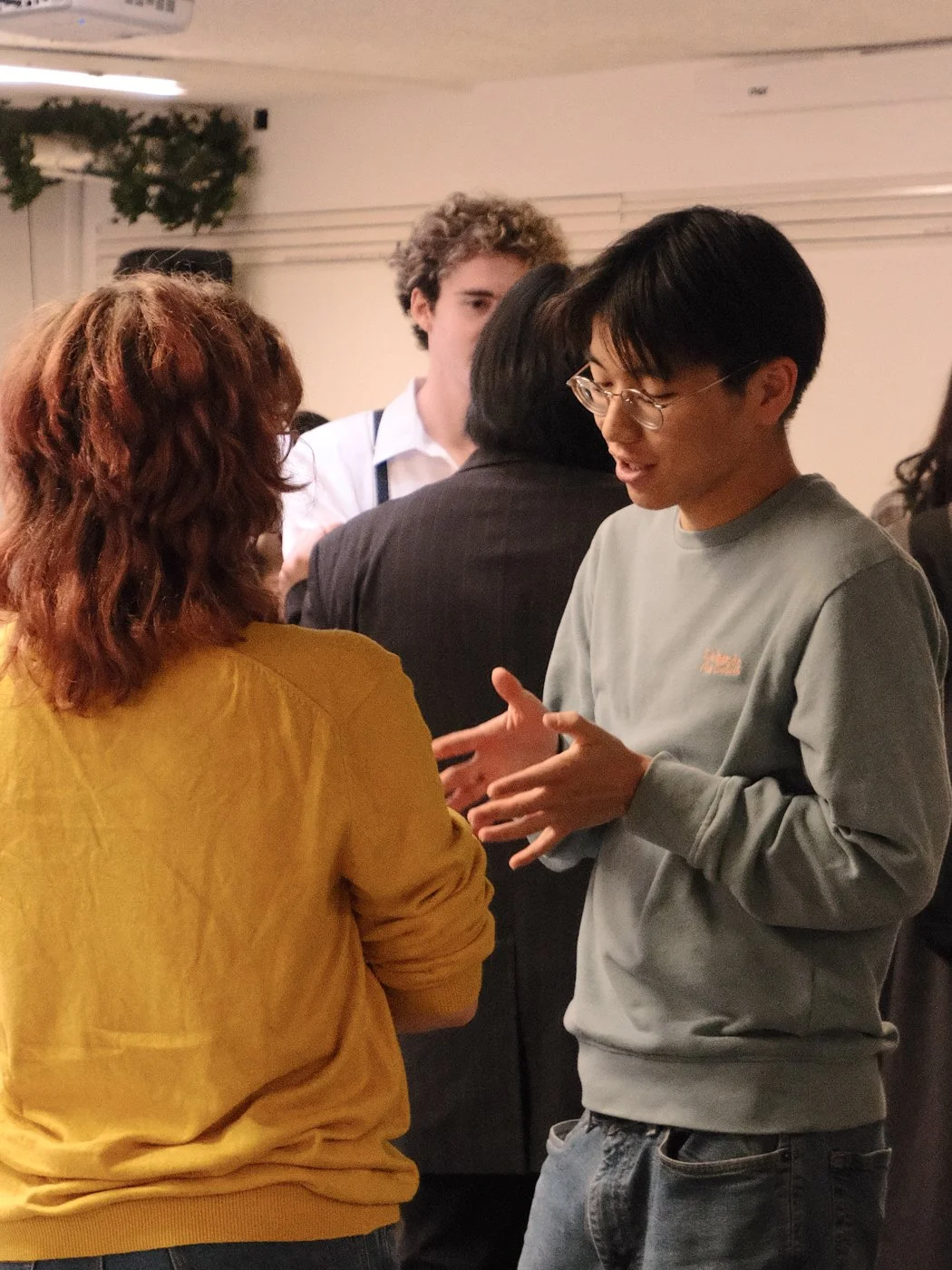 Deux jeunes personnes parlent lors d'une réunion ou d'un événement social, entourées d'autres personnes dans une salle. L'un porte un sweat gris et porte des lunettes, l'autre porte un pull jaune.