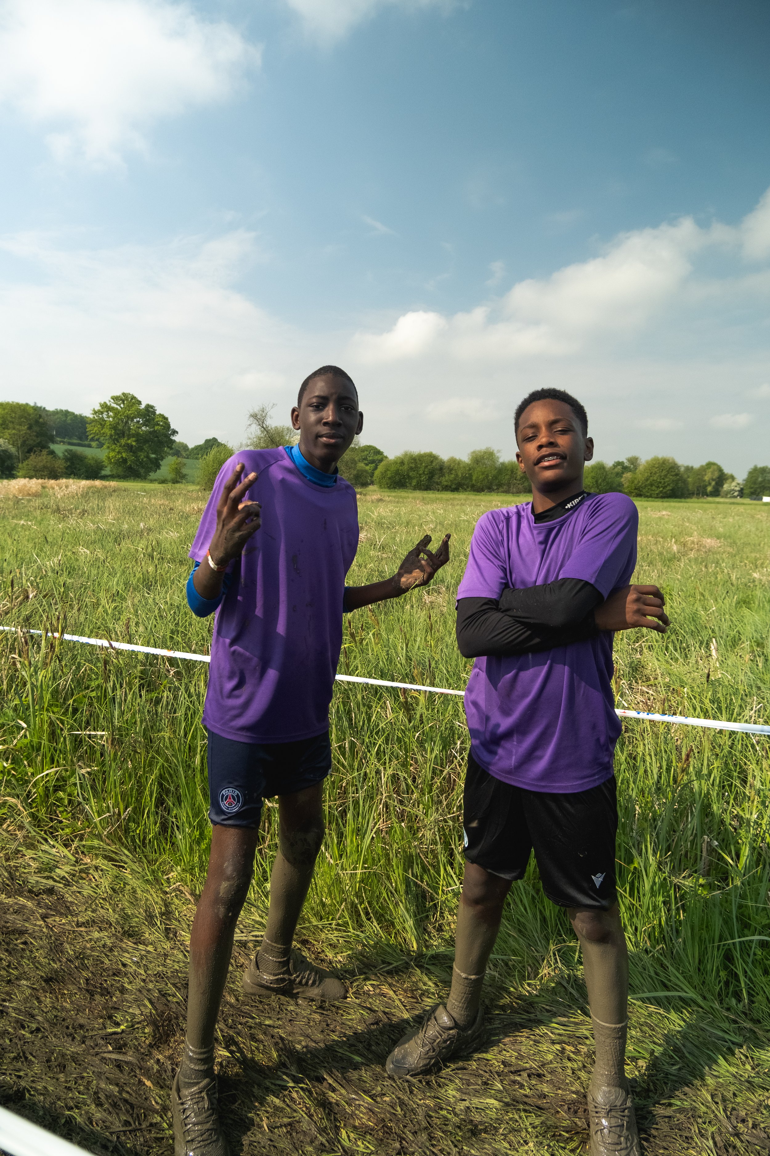Deux jeunes hommes en tenue de course, portant des vêtements violets, se tiennent dans un champ d'herbe verte sous un ciel partiellement nuageux.
