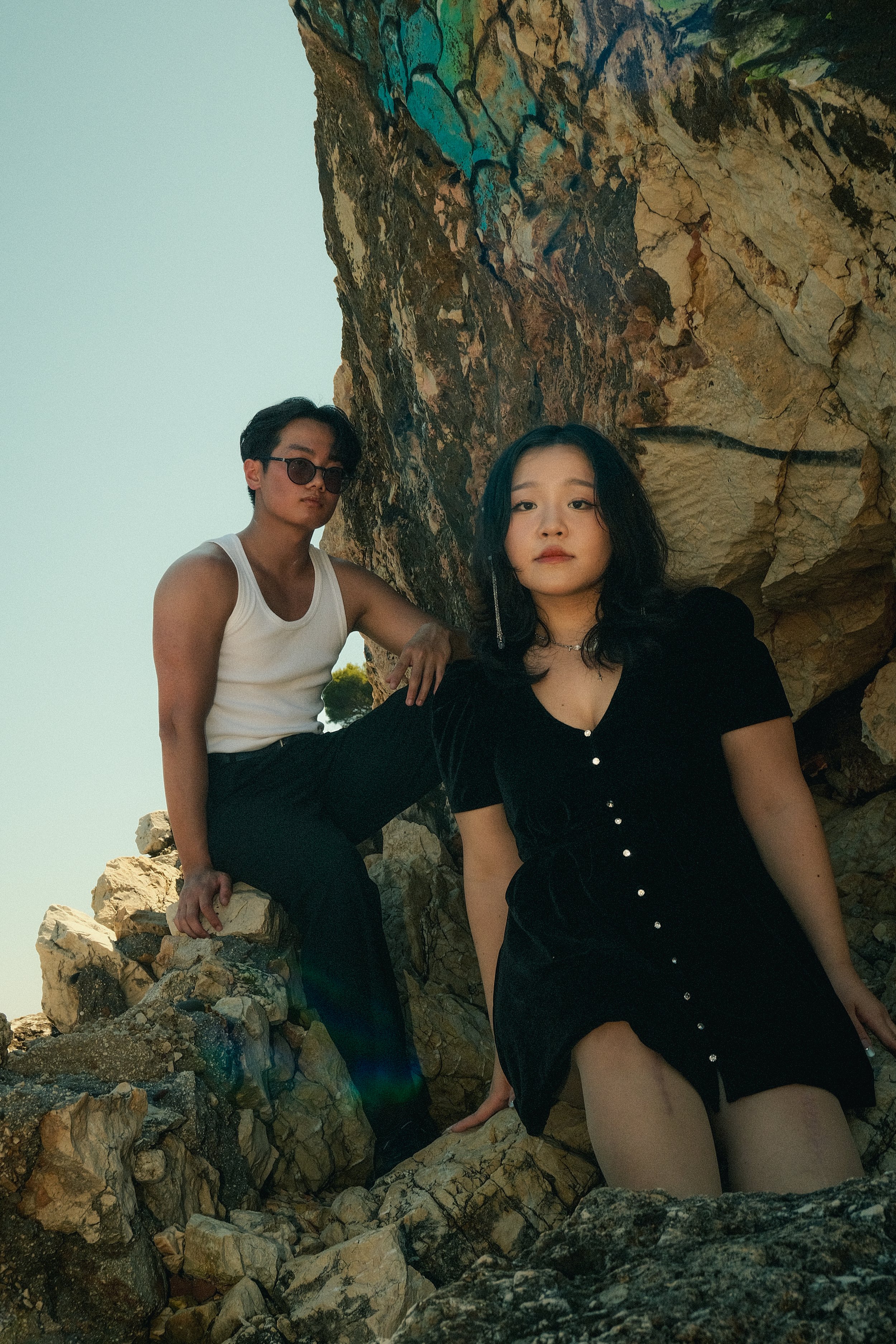 Deux jeunes adultes posent sur des rochers devant une falaise, vêtus de tenues décontractées, avec un ciel clair en arrière-plan.