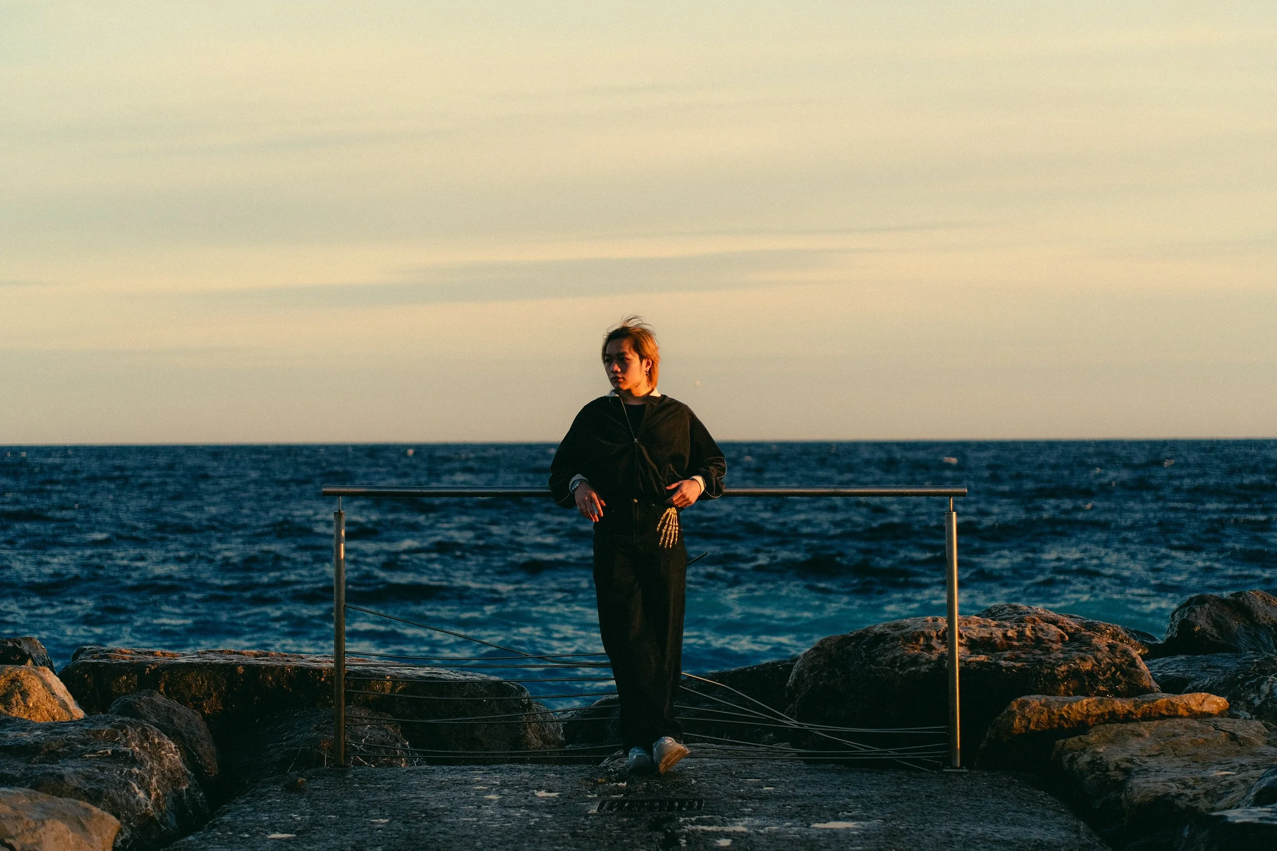 Une personne debout sur une jetée en pierre, au bord de la mer, au coucher du soleil, portant un sweat noir et un pantalon noir, avec une main dans sa poche et l'autre reposant sur la balustrade en métal.