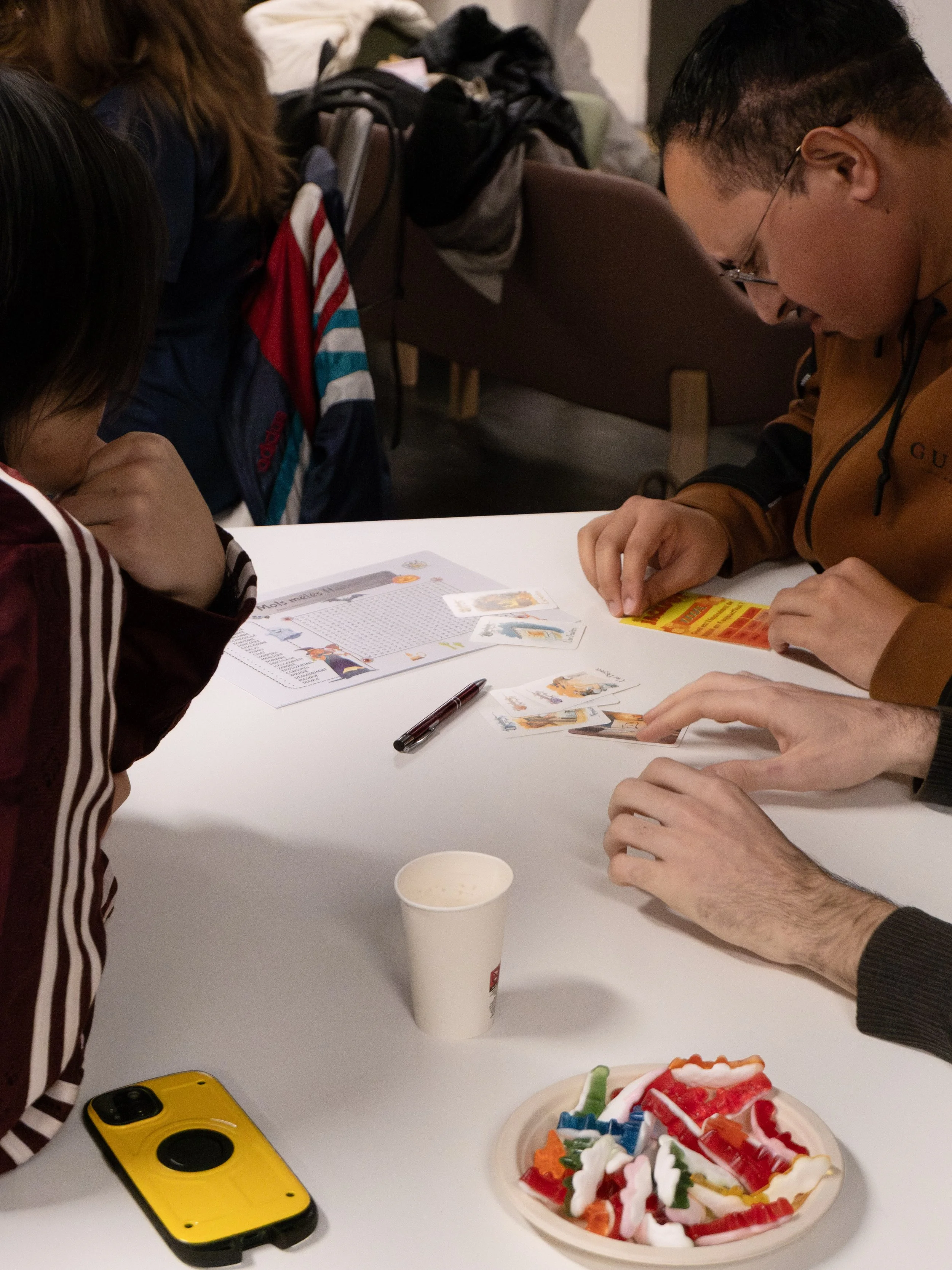 Groupe de personnes jouant à un jeu de cartes sur une table blanche, avec un smartphone jaune, une tasse en papier et un plateau de bonbons colorés.