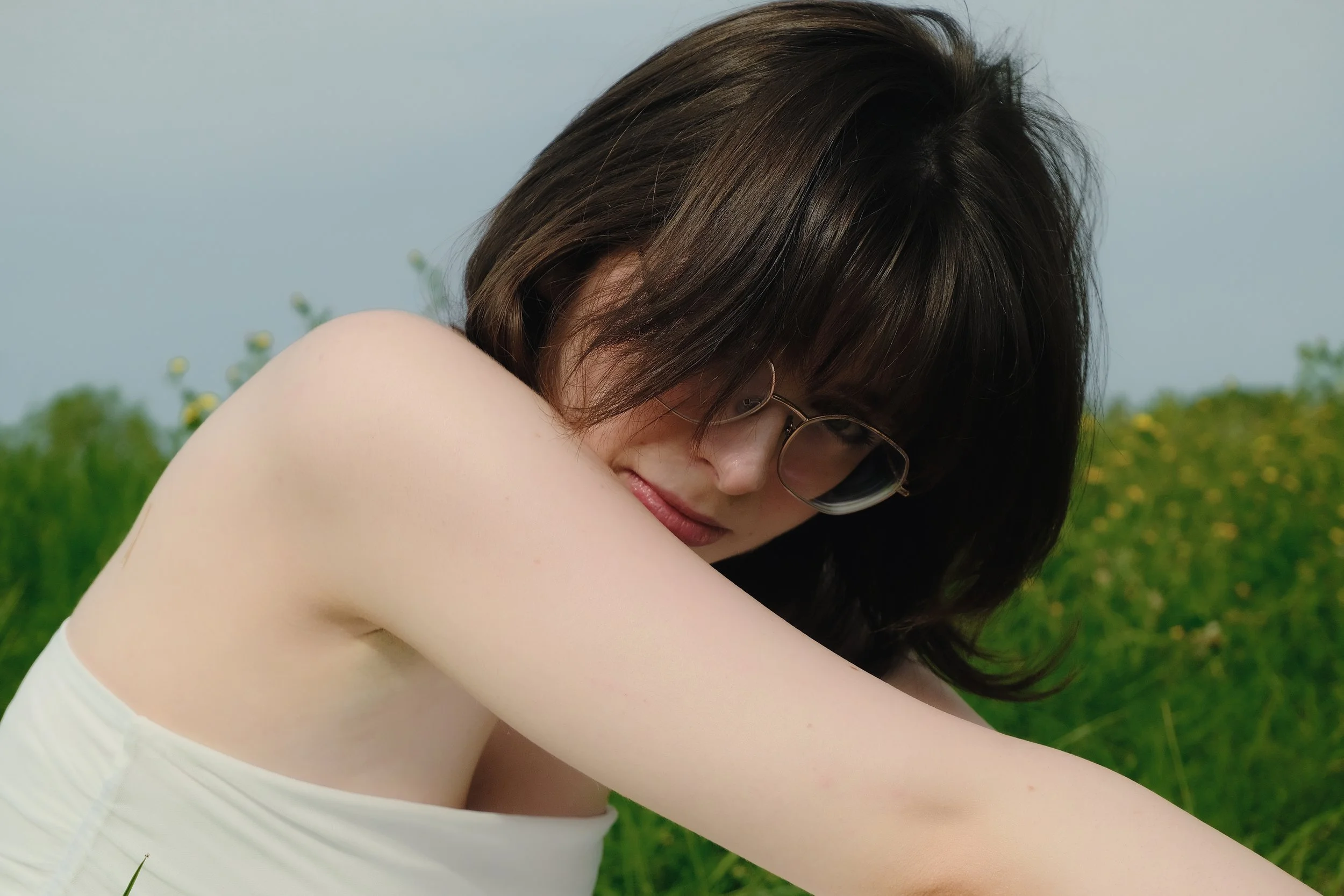 Jeune femme avec des lunettes, cheveux bruns, souriant doucement, dans un champs vert sous un ciel nuageux.