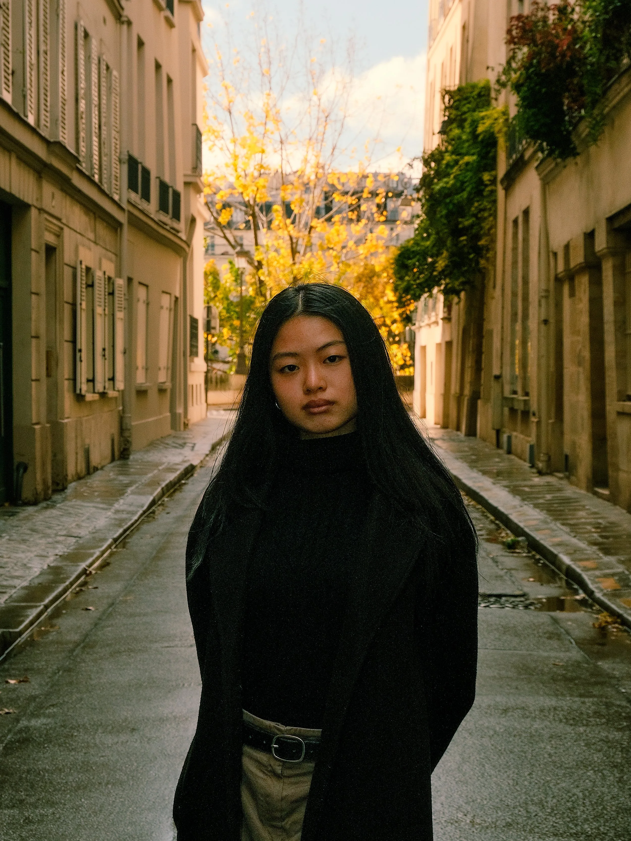 Jeune femme avec des cheveux noirs, portant un pull noir et un pantalon beige, se tenant dans une ruelle urbaine pavée avec des bâtiments anciens, arbres aux feuilles jaunes en arrière-plan, ciel partiellement nuageux.