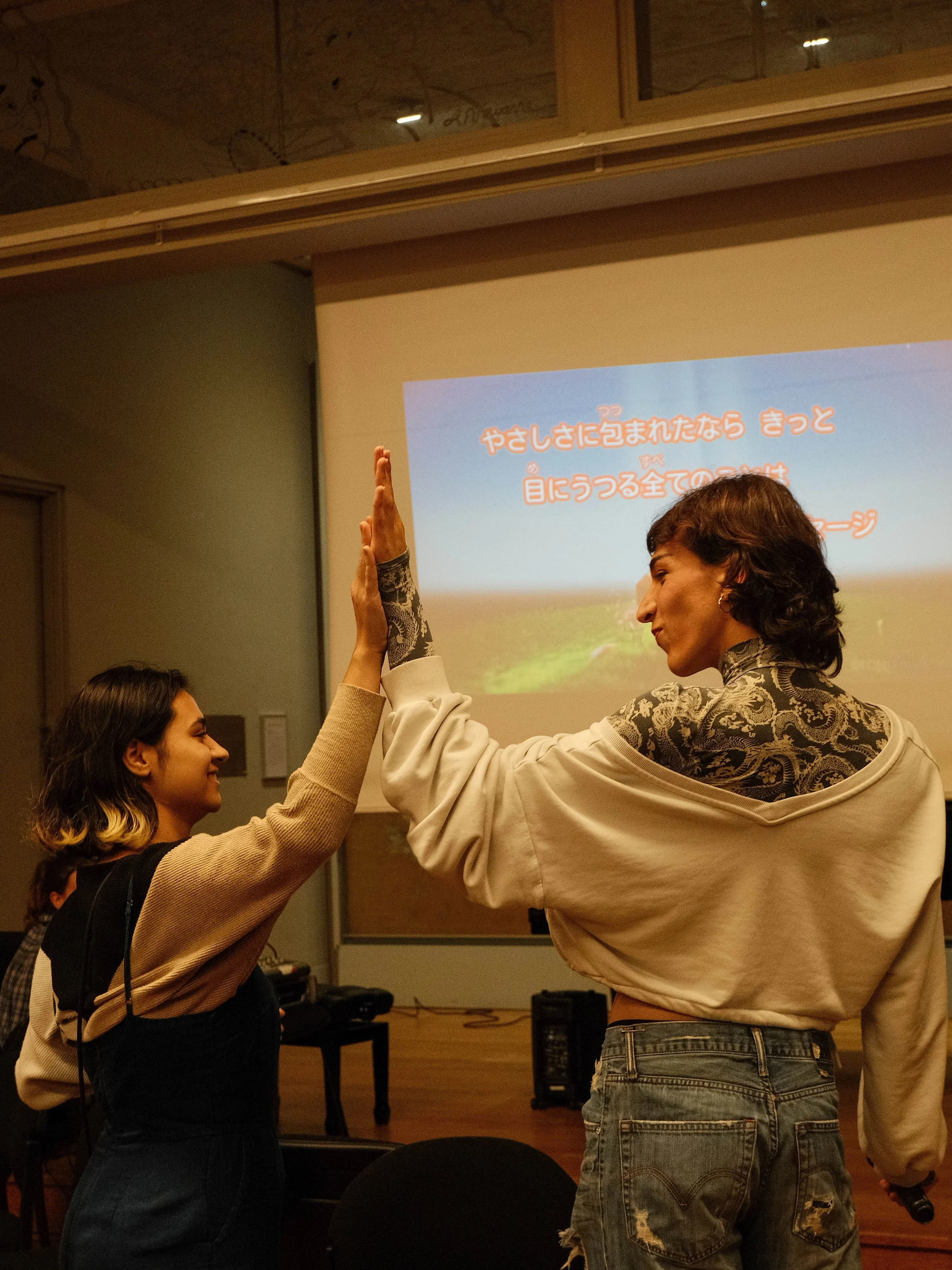 Deux jeunes femmes se donnent un high-five en face d'un écran de projection affichant un texte en japonais, dans une pièce intérieure.