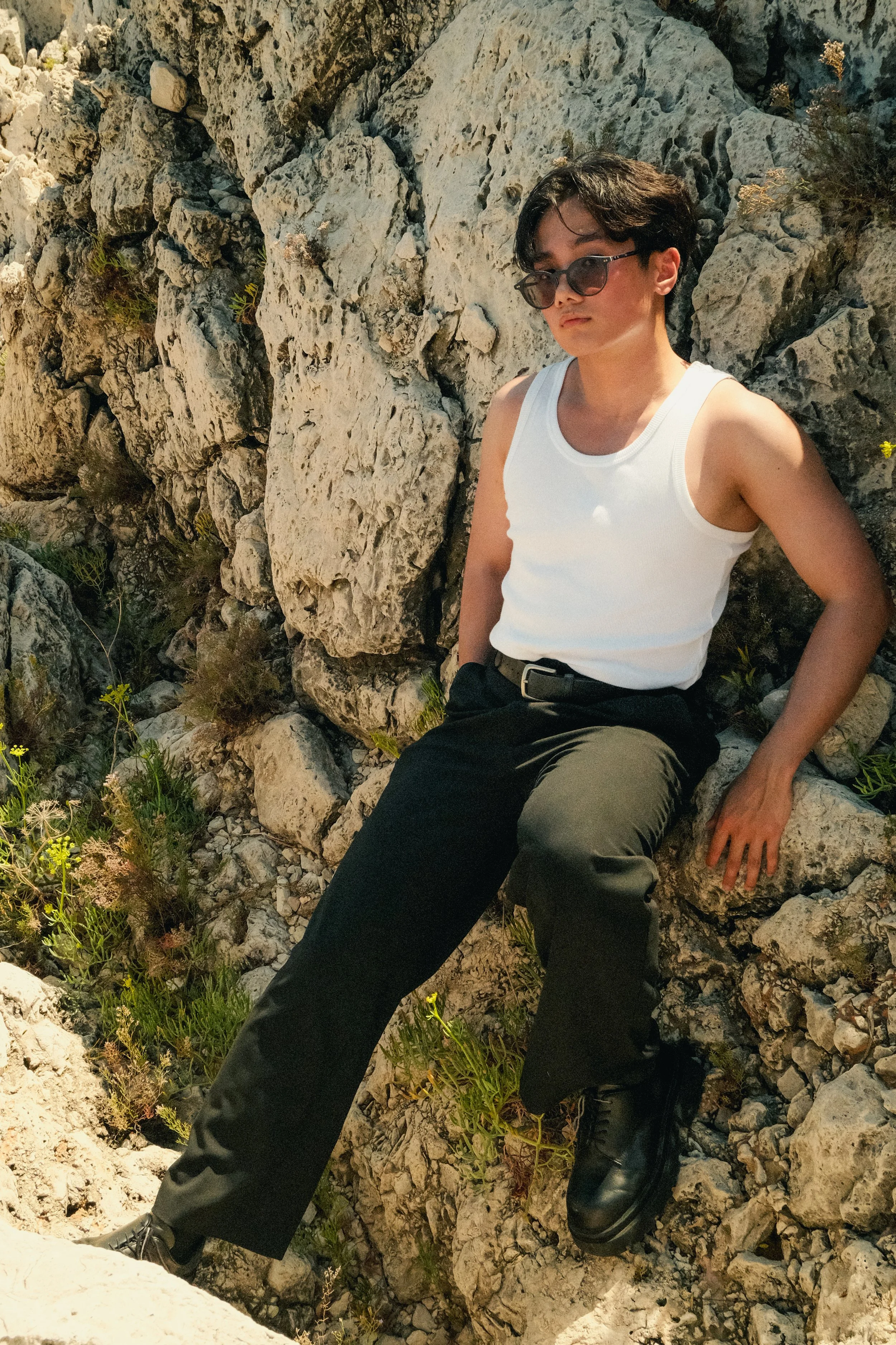 Jeune homme assis contre des rochers, portant un débardeur blanc, des pantalons noirs, des lunettes de soleil et des bottes noires, dans un environnement rocheux.