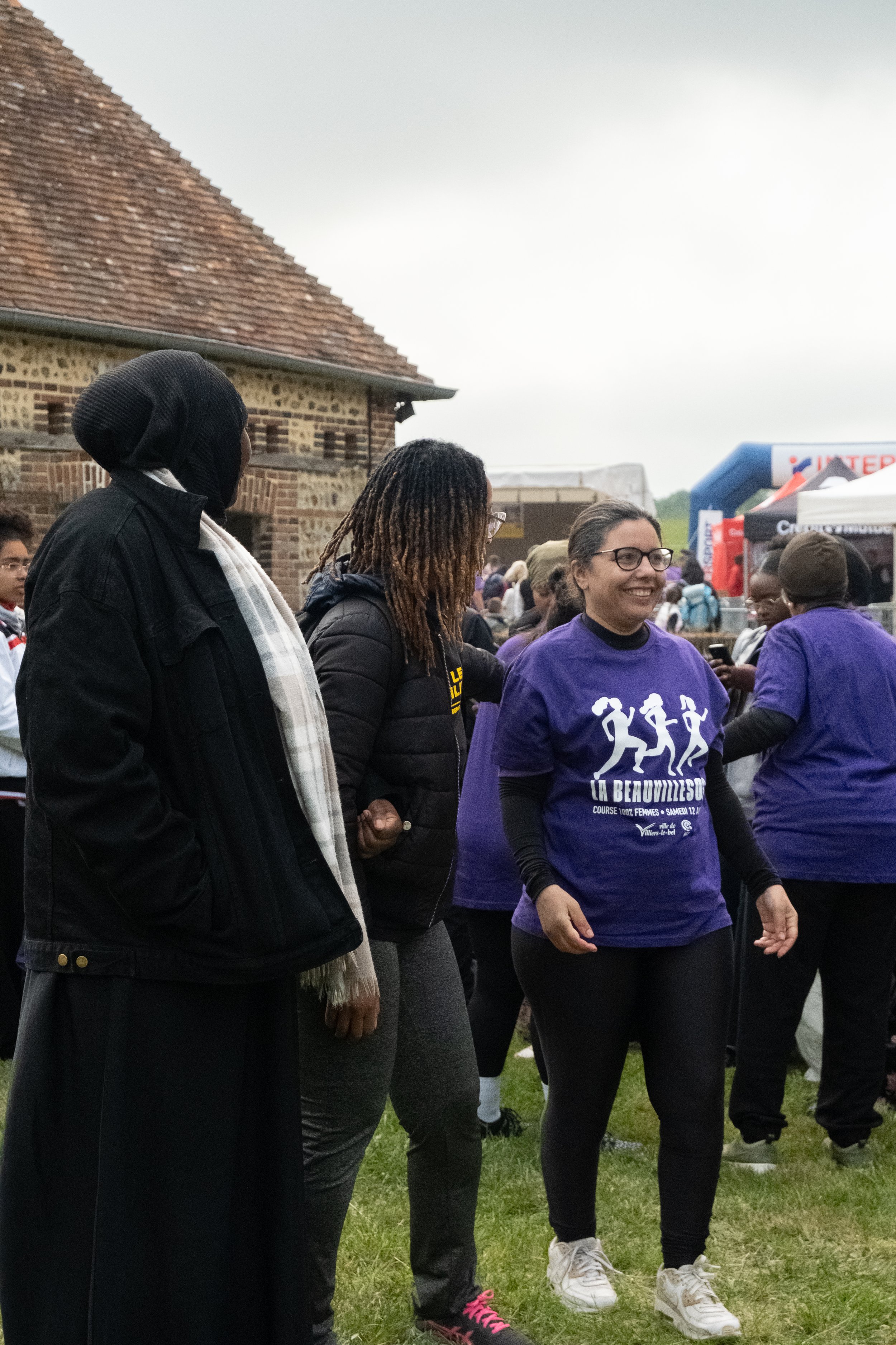 Groupe de femmes participant à une course organisée, portant un t-shirt violet avec un dessin de femmes courant et le texte « La Beauvilloise ». Le rassemblement se déroule à l'extérieur, avec des bâtiments en arrière-plan et d'autres personnes prése