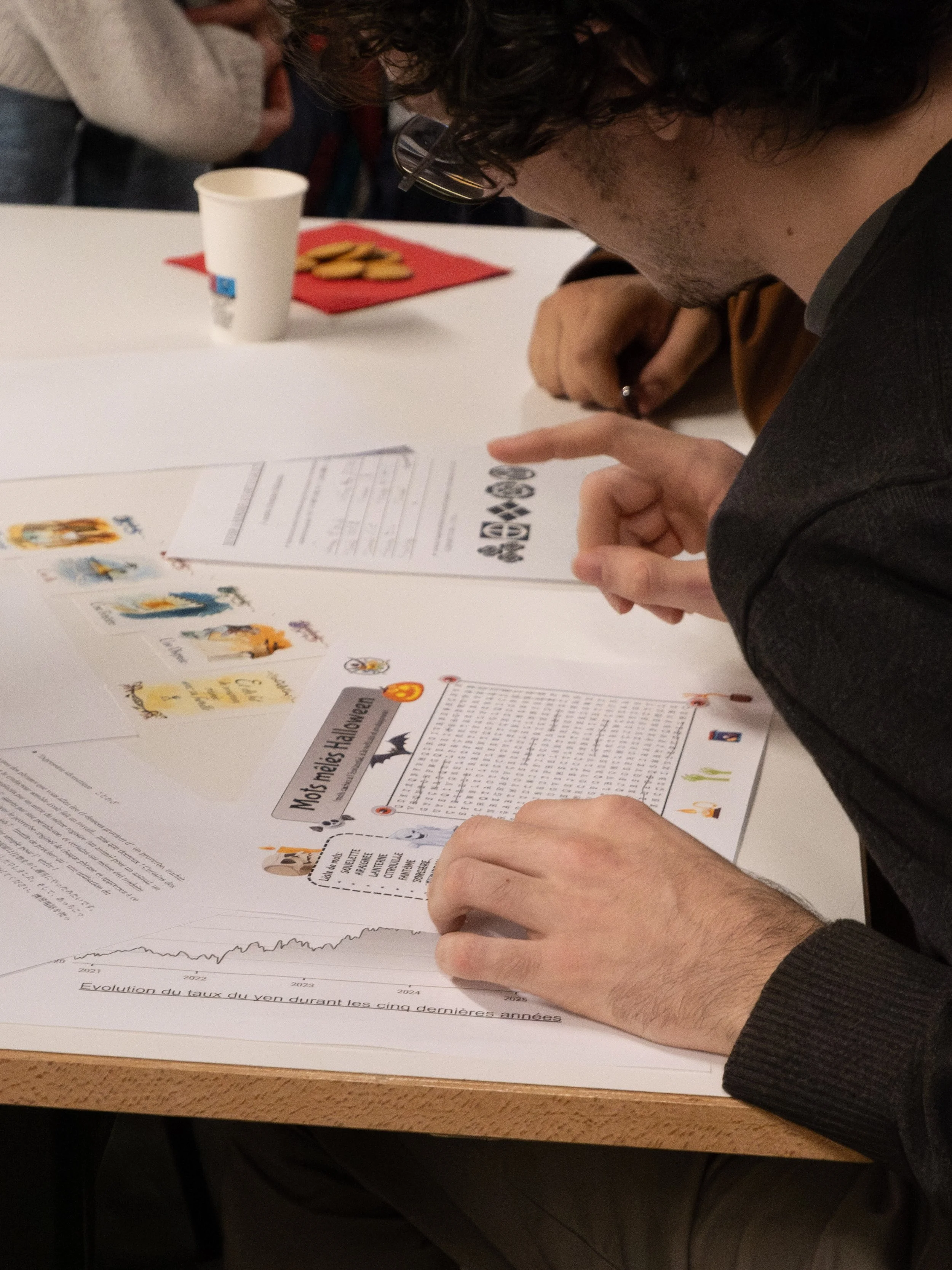 Une personne à lunettes qui lit un document, avec des papiers et des illustrations sur la table, dans un environnement de bureau ou de réunion.