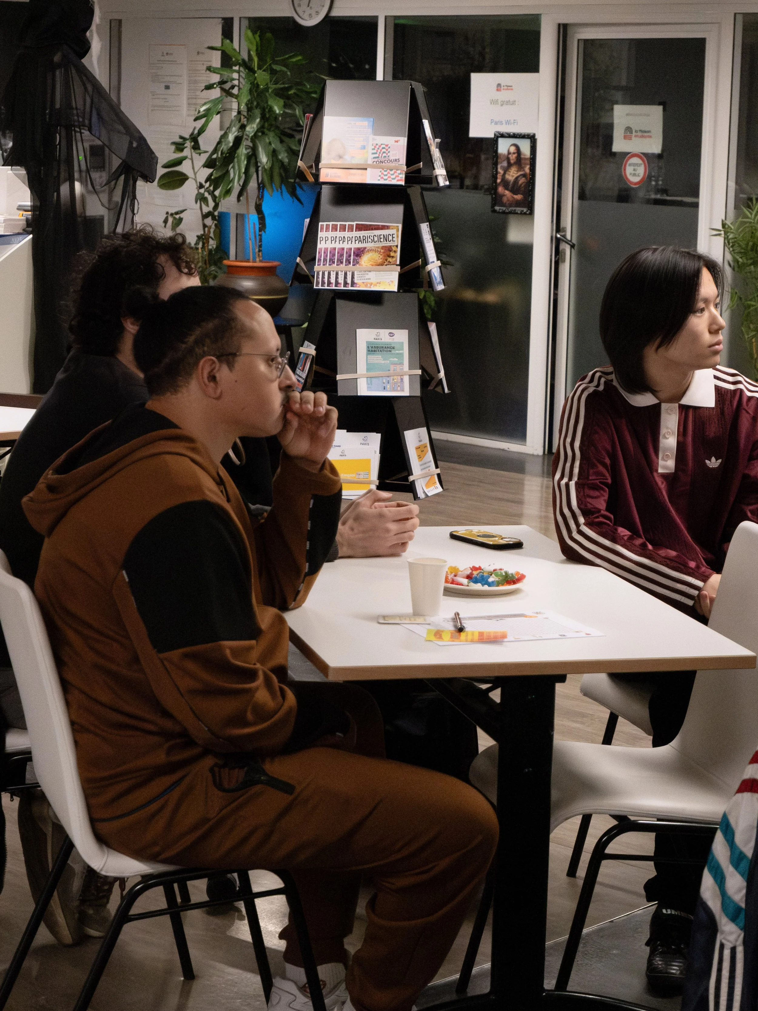 Groupe de personnes assises autour d'une table lors d'une réunion ou d'un atelier, avec des brochures et des collations. Décor intérieur avec plantes et étagère.