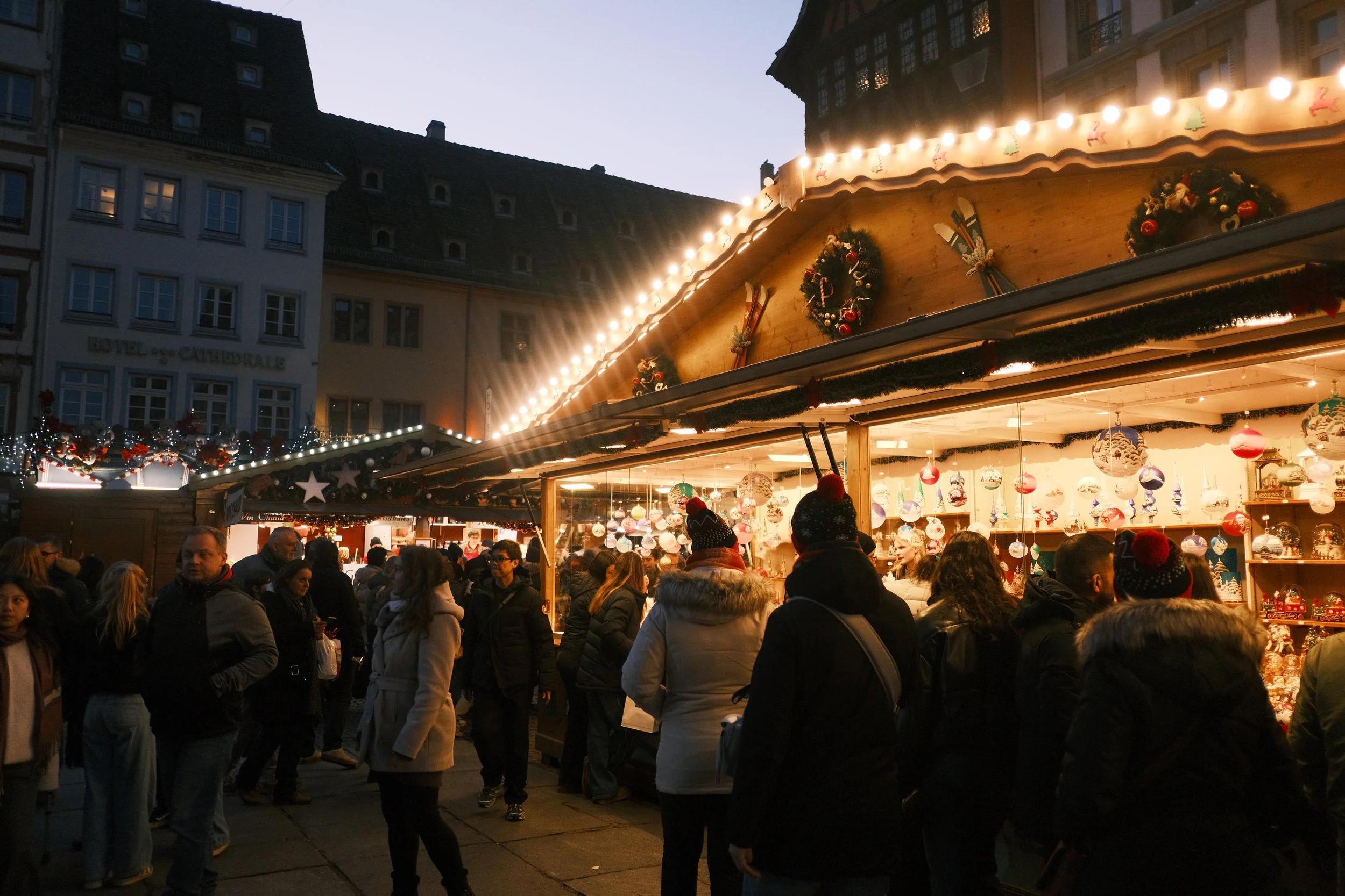 Marché de Noël avec des stands décorés de guirlandes lumineuses, des gens faisant du shopping, et des décorations de Noël suspendues.