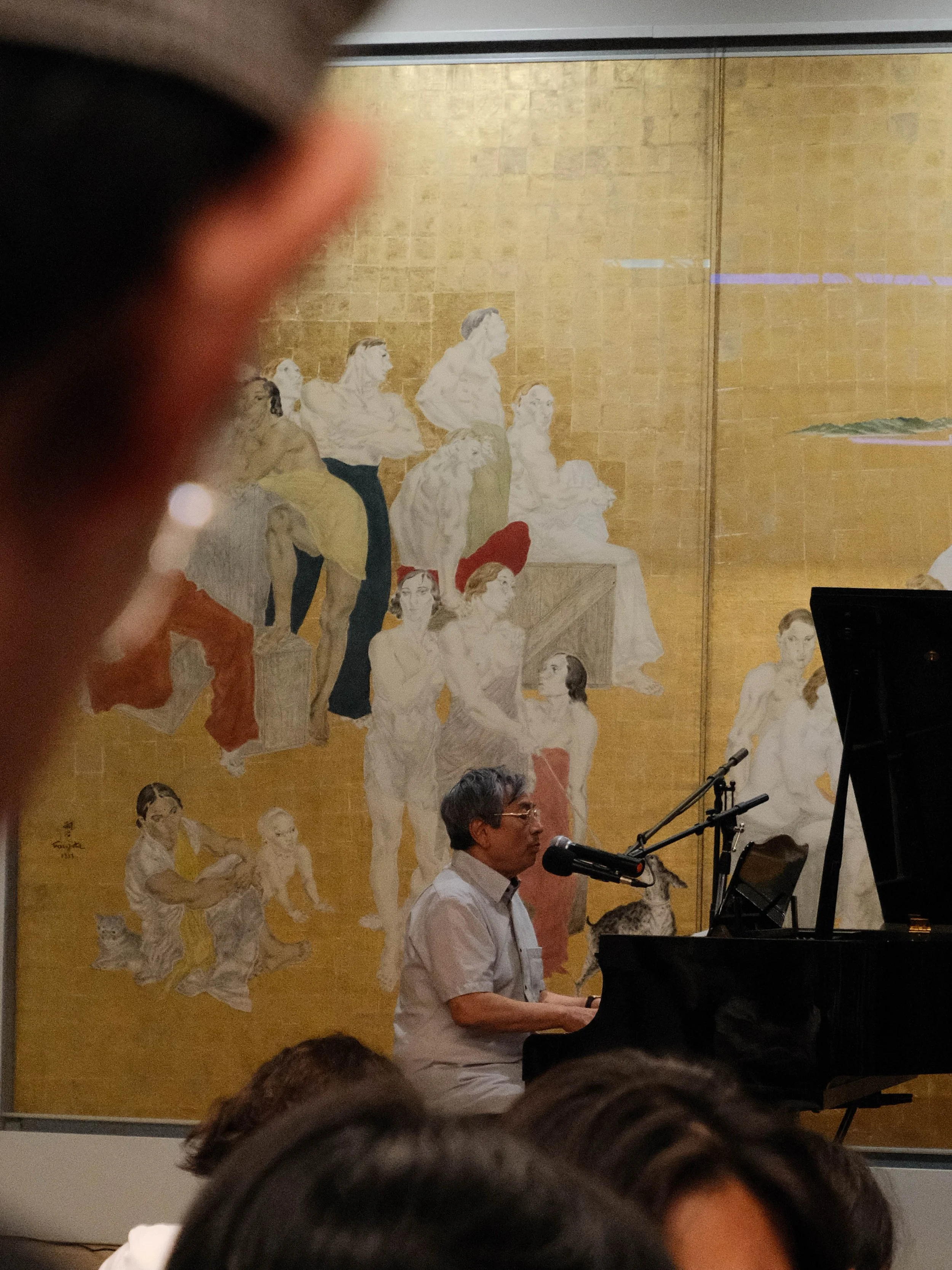 Un homme joue du piano et chante devant une grande fresque murale illustrant plusieurs figures humaines, dans un lieu intérieur.