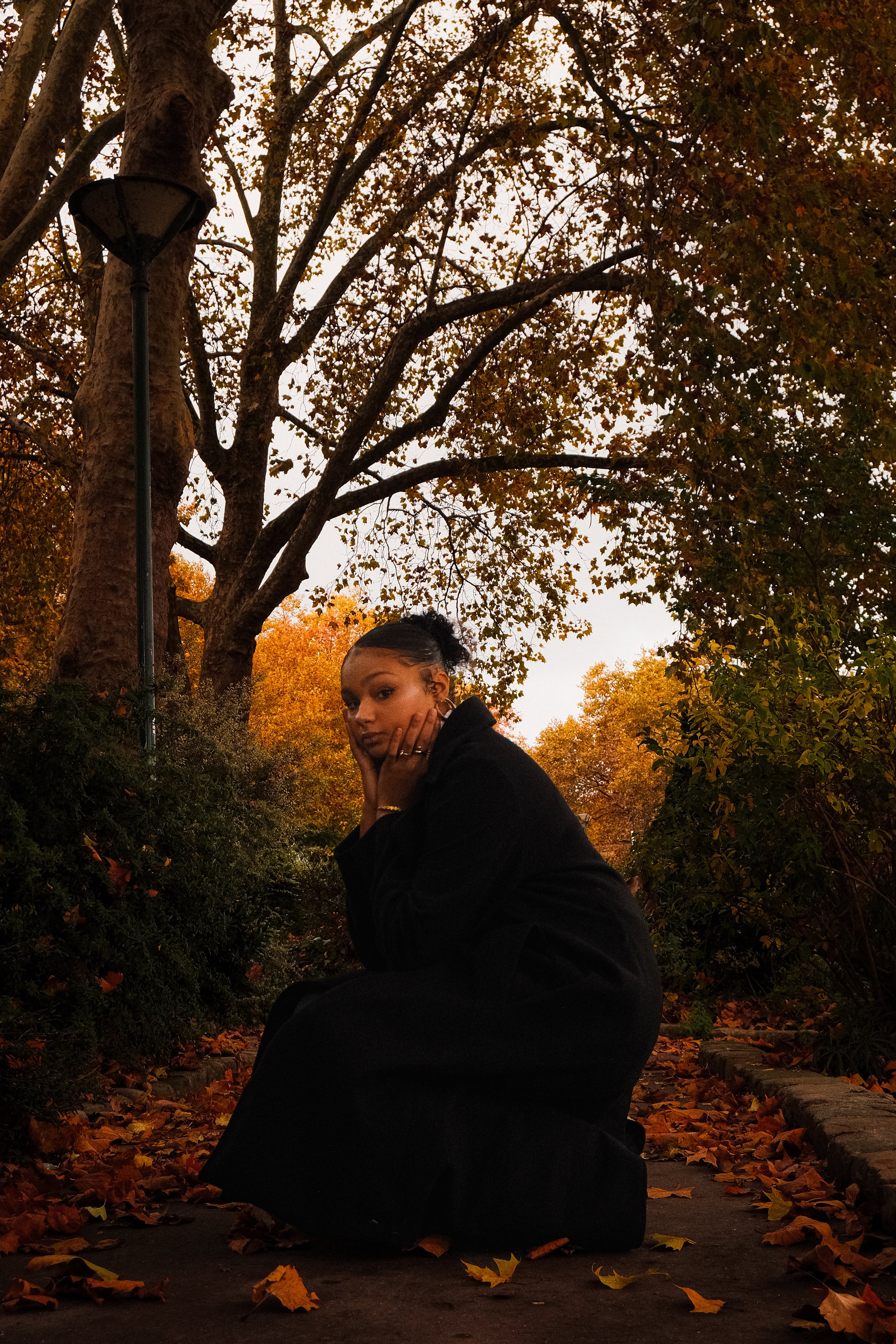 Une femme assise sur un sentier bordé d'arbustes et de feuilles d'automne, portant un manteau noir, sous un arbre aux feuilles orangées.