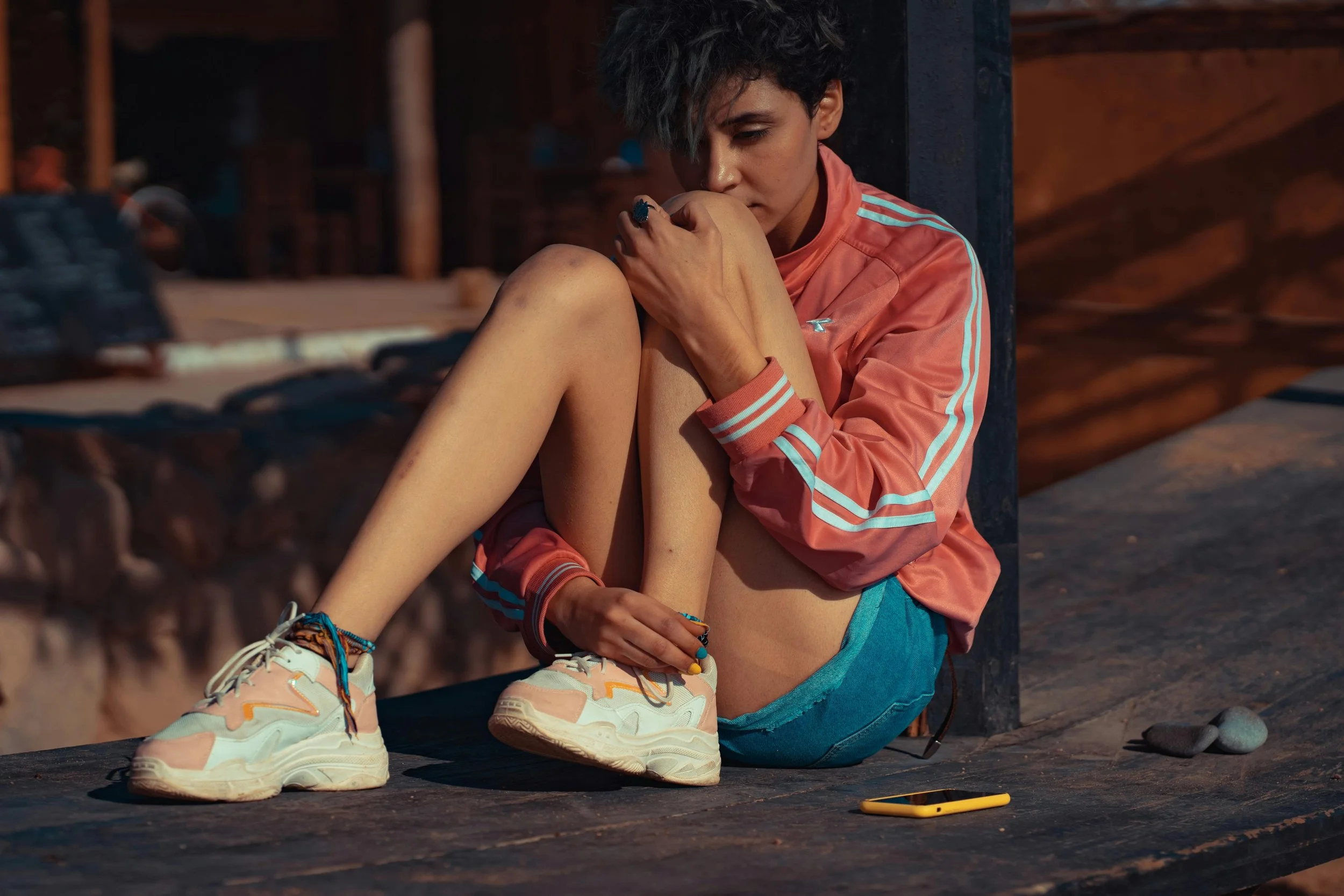 A young woman with short dark hair, wearing a peach-colored jacket and denim shorts, sitting on a wooden platform, holding her knee close to her face, with a pensive expression.