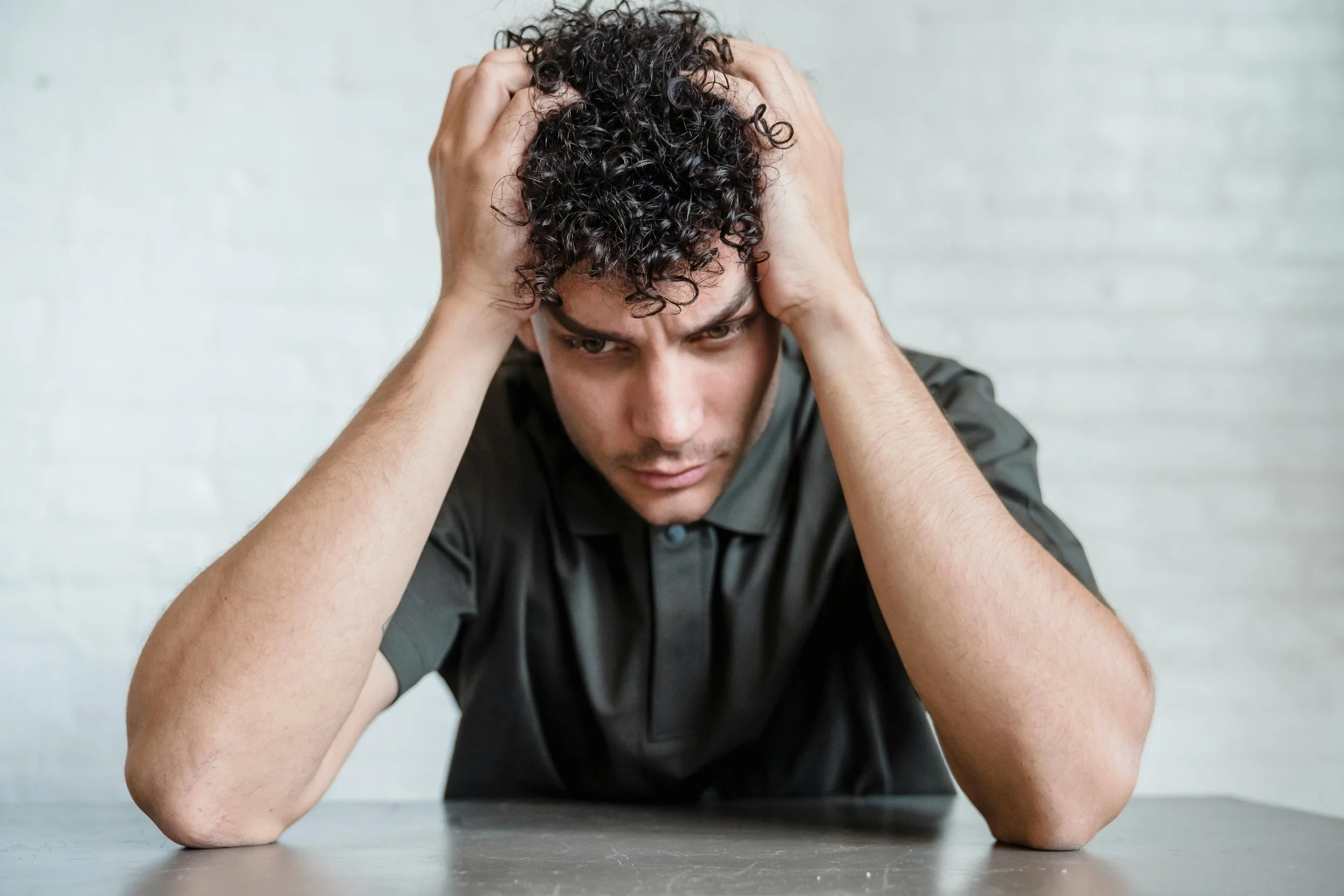 A young man sitting at a table, holding his head and looking downward with a distressed expression.