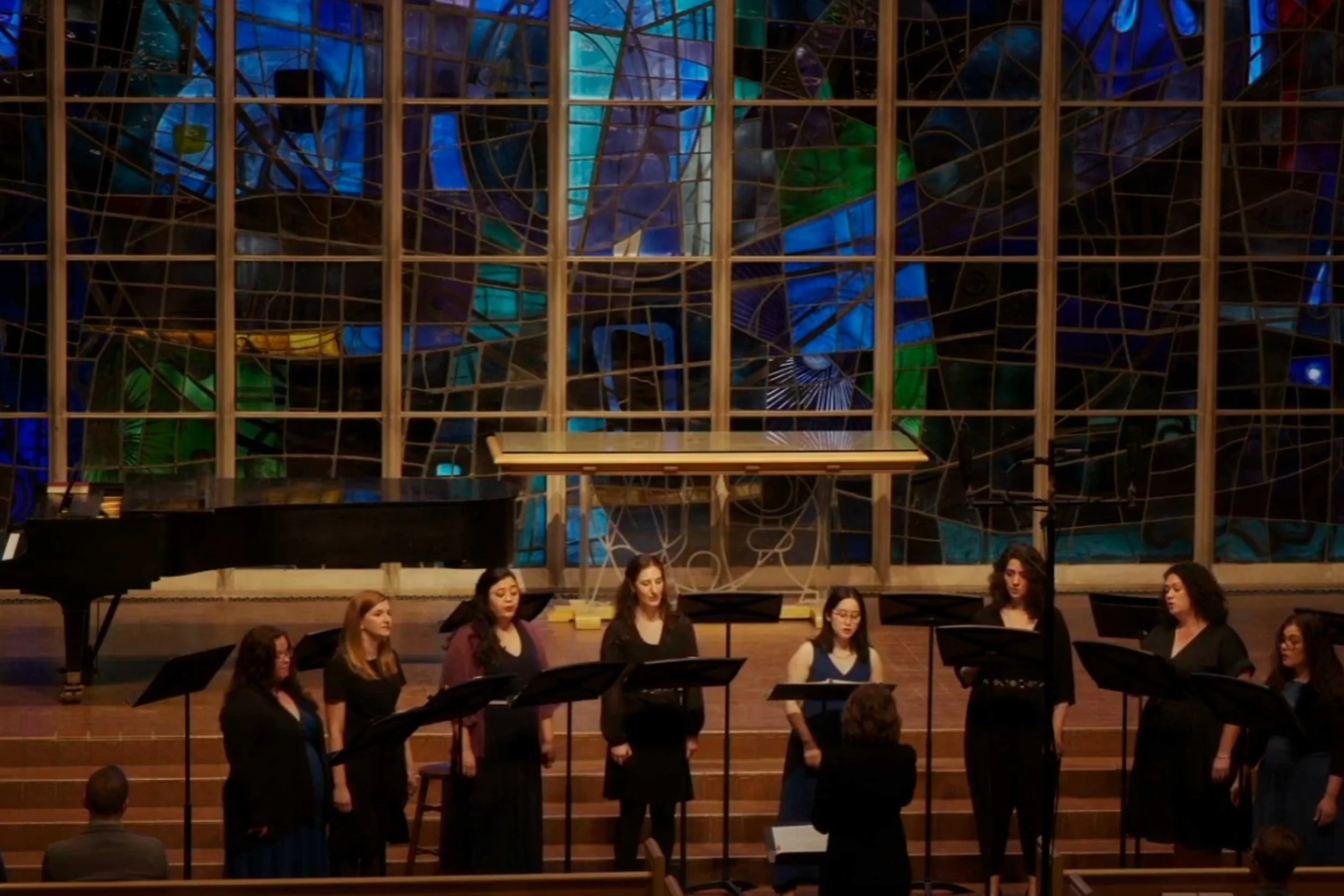 A choir of women singing on stage in front of a stained glass window in a church or concert hall, with a grand piano on the left side and a conductor guiding them.