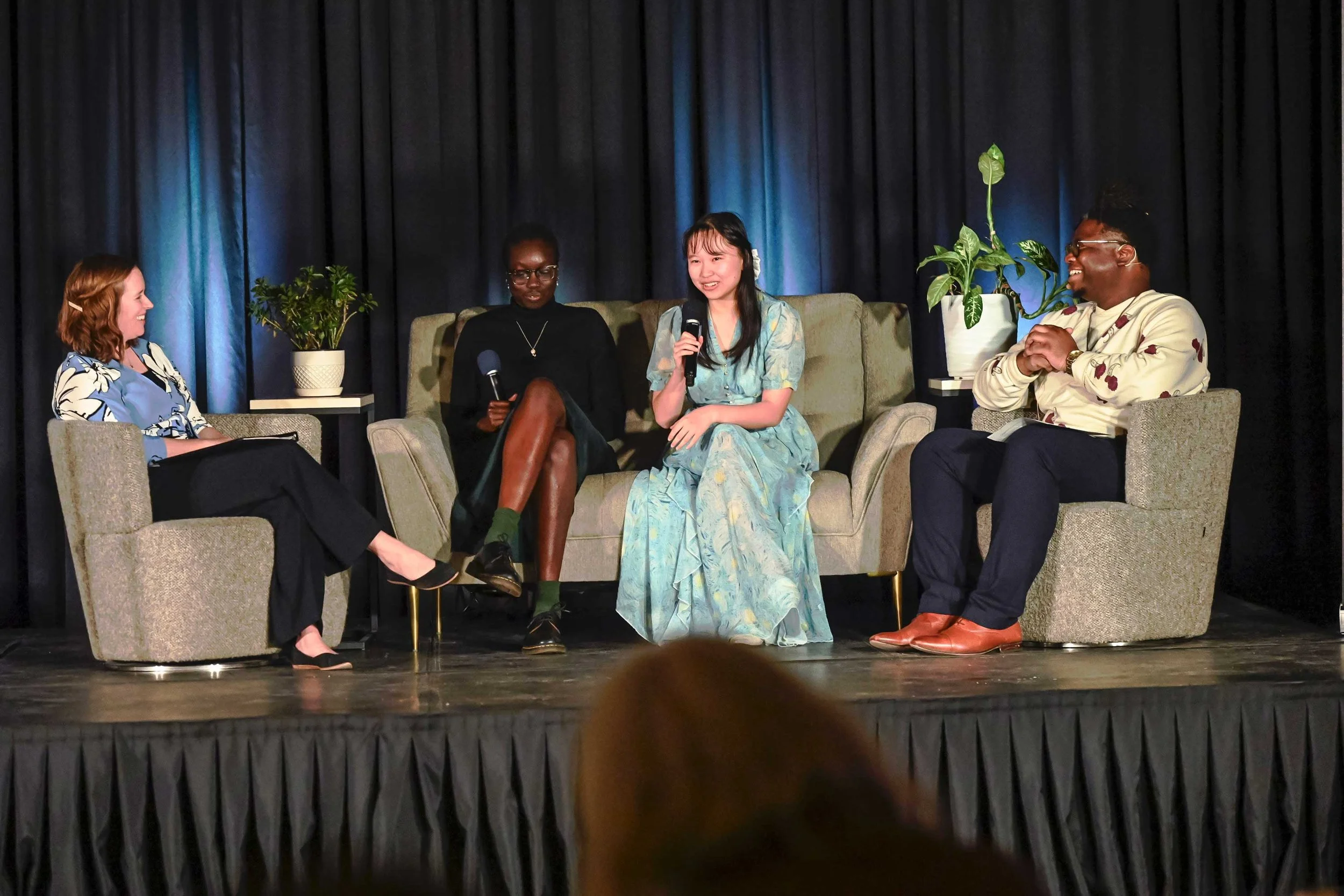 Four women sitting on a stage in a panel discussion, with two of them holding microphones. There are potted plants on side tables, and a dark curtain backdrop.