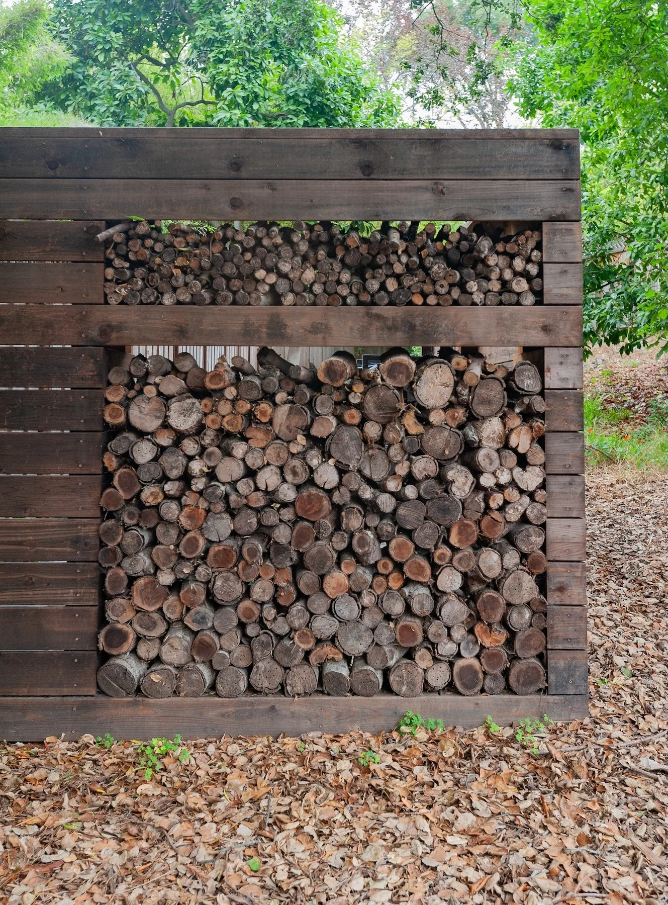 Wooden firewood storage rack filled with stacked logs, set in a gravel garden bed - Orchard Keepers