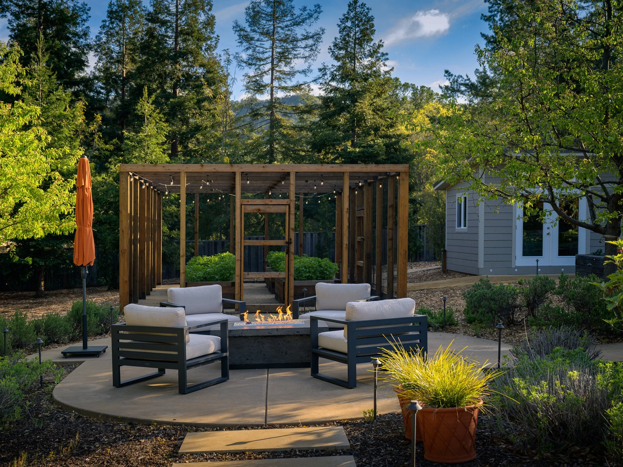 A backyard with a fire pit, surrounded by outdoor furniture including cushioned chairs, trees, a wooden pergola, and a house in the background on a clear day.