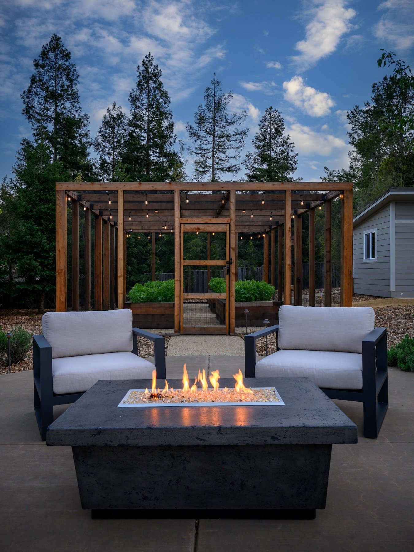 A backyard patio at dusk with a fire pit, two white cushioned chairs, and a wooden pergola with string lights, surrounded by trees and a small house on the right.