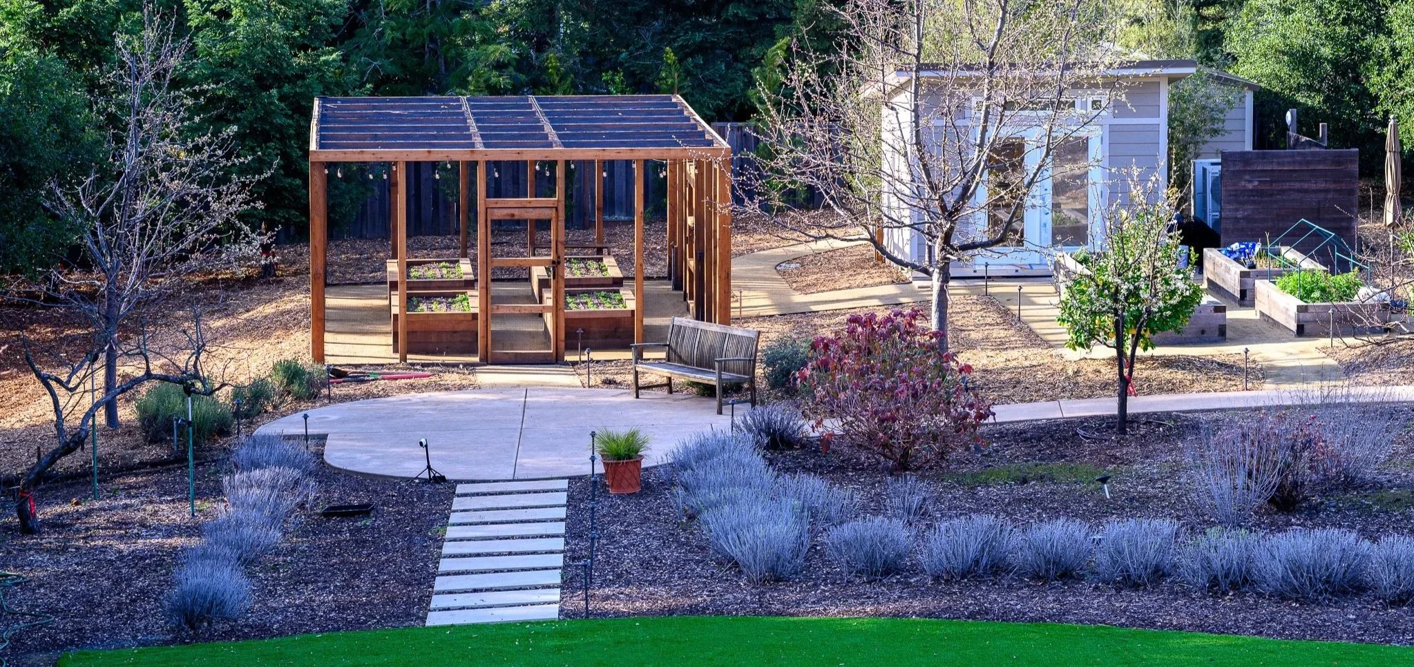 A backyard garden features a small wooden greenhouse, a raised planting bed, a garden shed, trees, shrubs, and a bench, with a curved pathway and various plants and flowers.