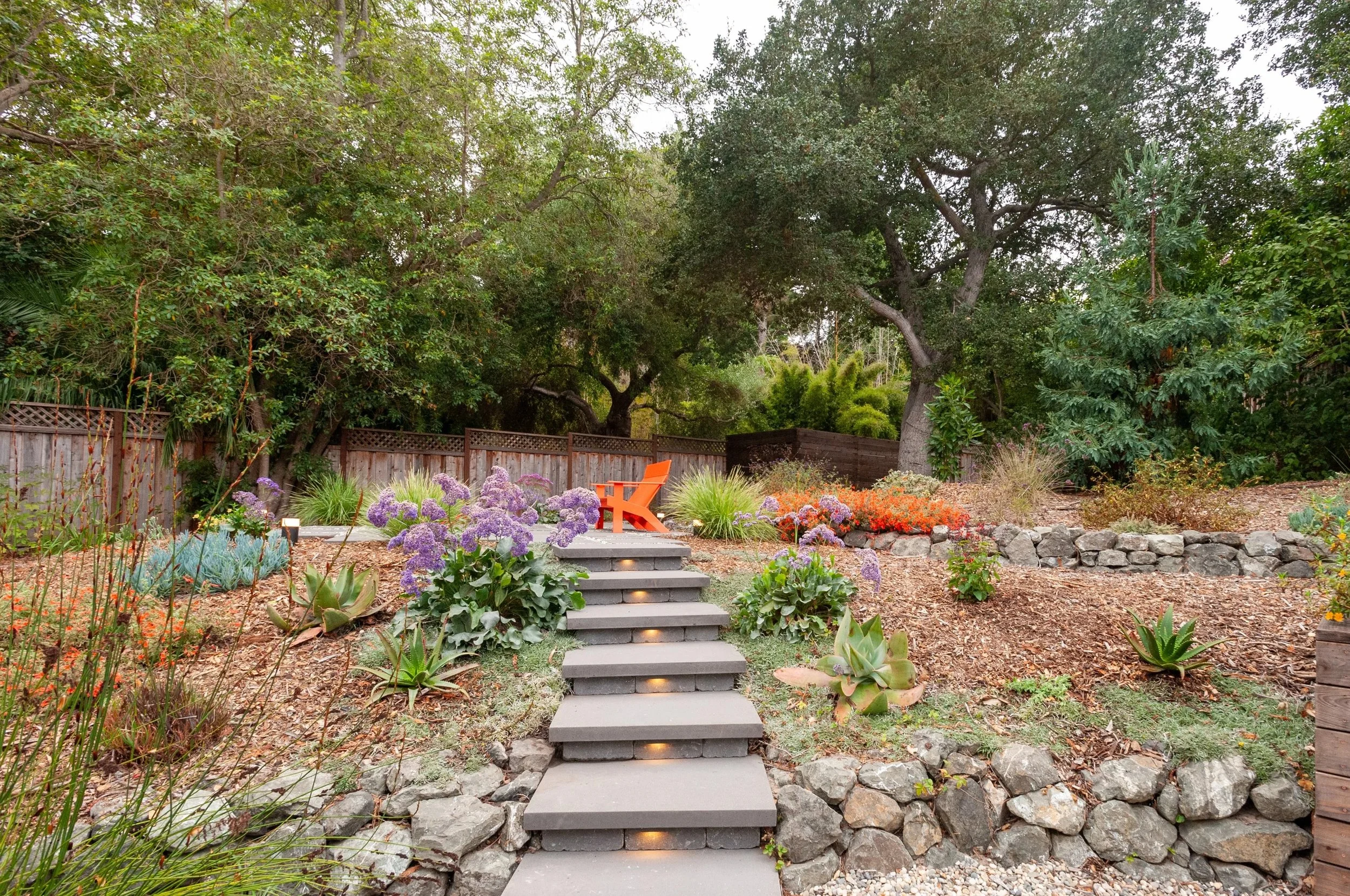 A garden with steps leading up to a sitting area with orange chairs, surrounded by various plants and flowers, with large trees in the background and a wooden fence.