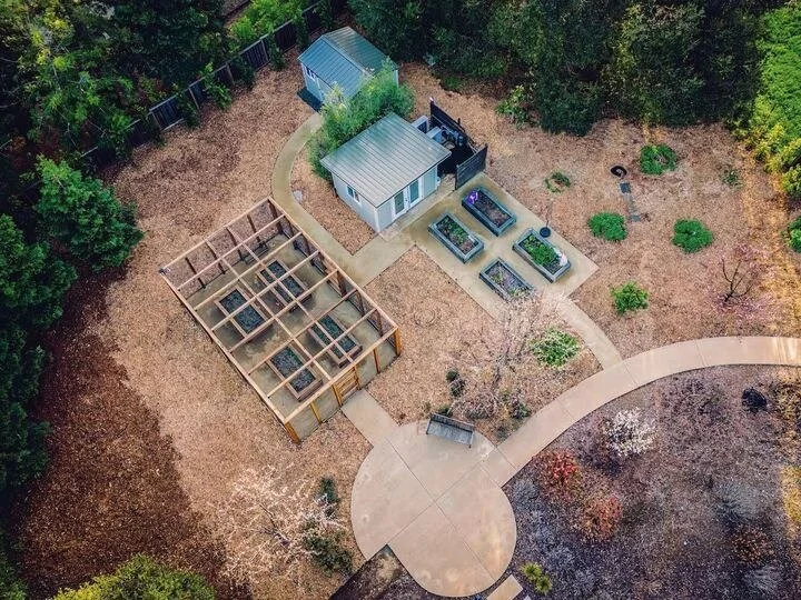 Aerial view of a backyard with a small house, a shed, garden beds, and a wooden chicken coop, surrounded by trees and a circular concrete path.