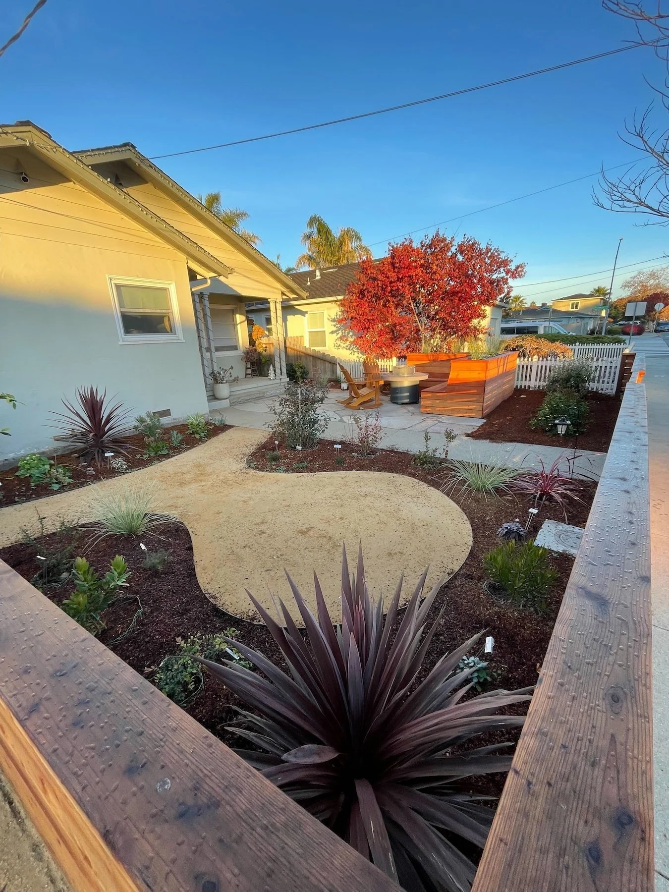 View of a backyard garden with a circular sandy area, plants, a potted plant near a front house steps, a red tree, and a wooden planter with a chair and small table, under a clear blue sky.
