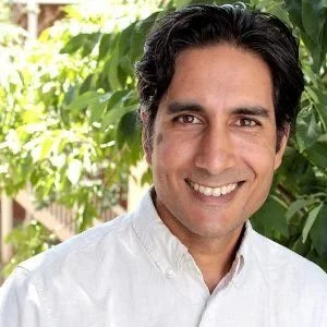 A man with dark hair wearing a white shirt, smiling outdoors with green foliage in the background.