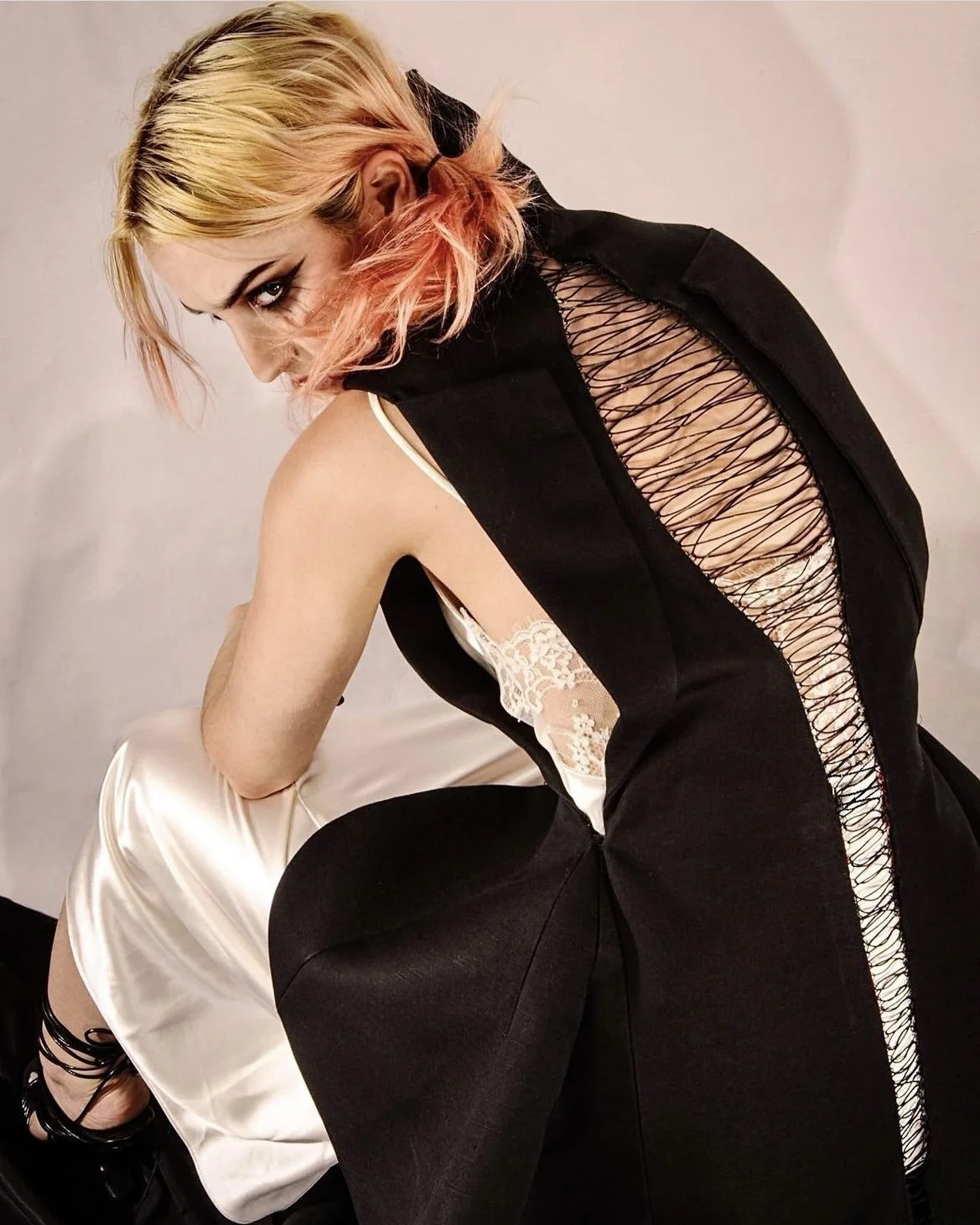 A woman with pink and blonde hair leaning over, wearing a black dress with a lace and string back detail and sitting on a person's lap in a white satin slip dress.