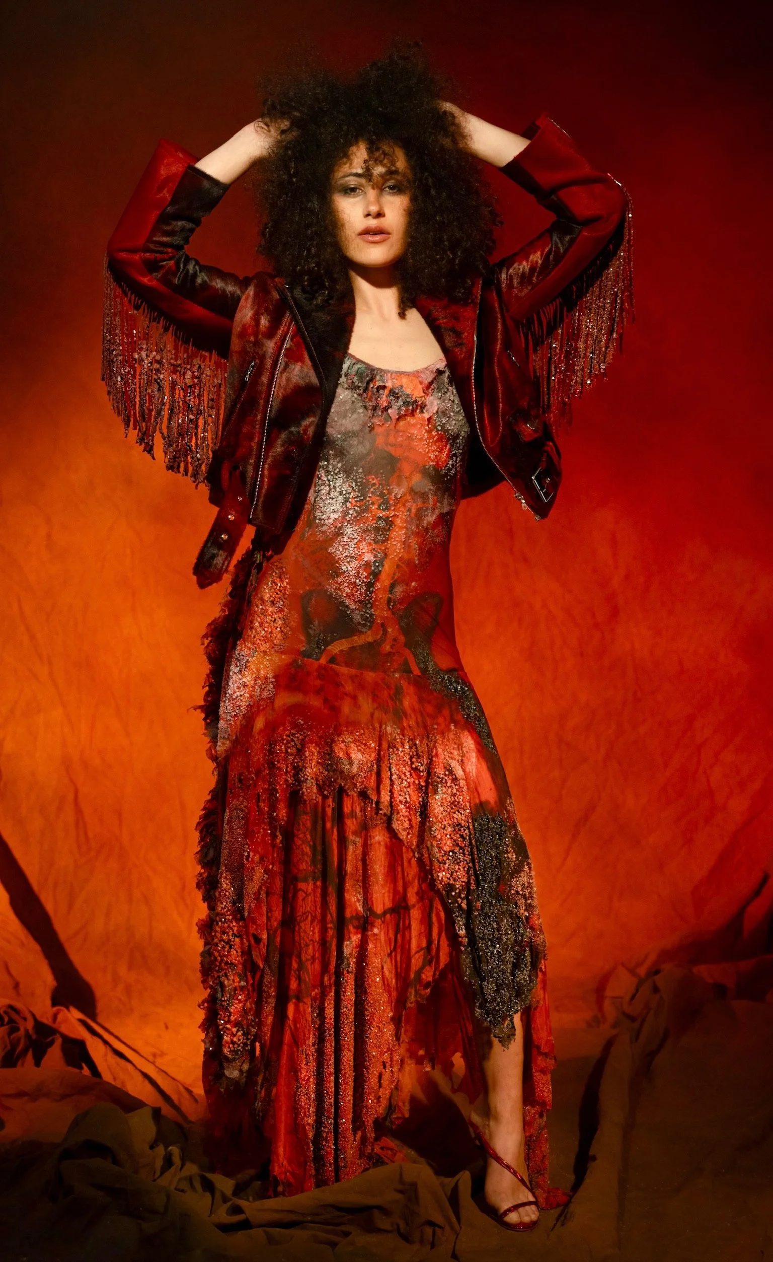 A woman with curly hair wearing a red and black outfit with a fringed leather jacket and a colorful, abstract dress standing against a red background.