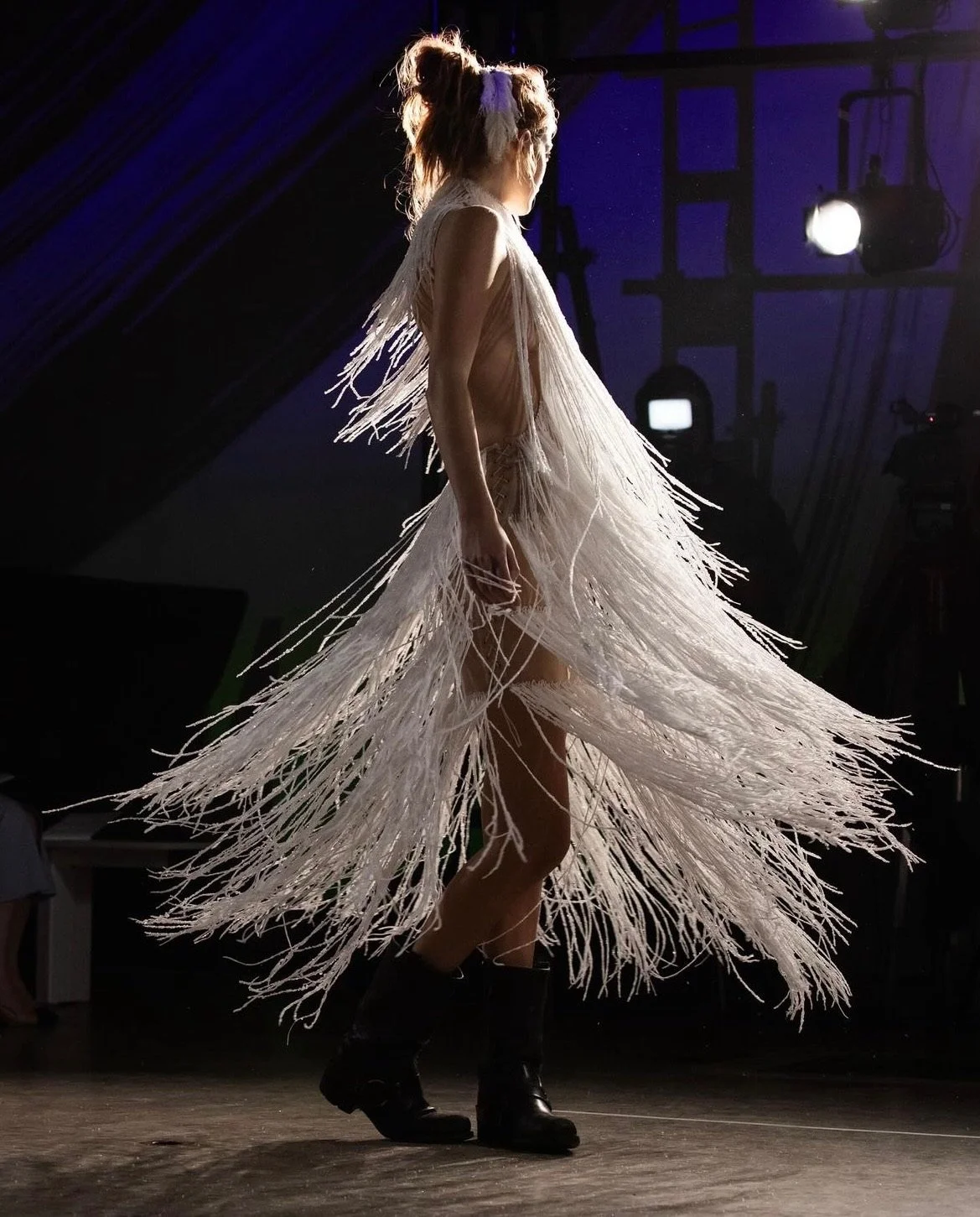 A female model walking on a runway wearing a long, fringed dress made of light-colored, wispy material with an edgy style, paired with black boots.