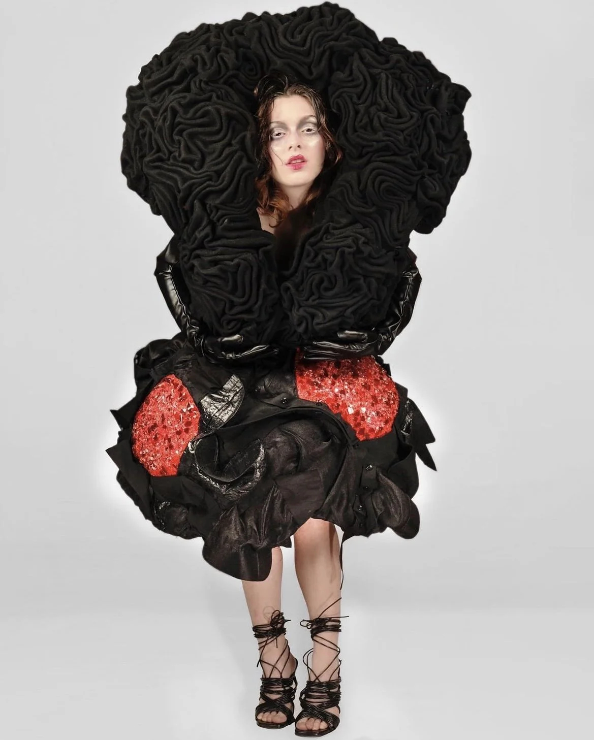 Fashion model wearing a large black ruffled garment with red sequined patches, tall black lace-up heels, and black gloves, posing against a plain background.
