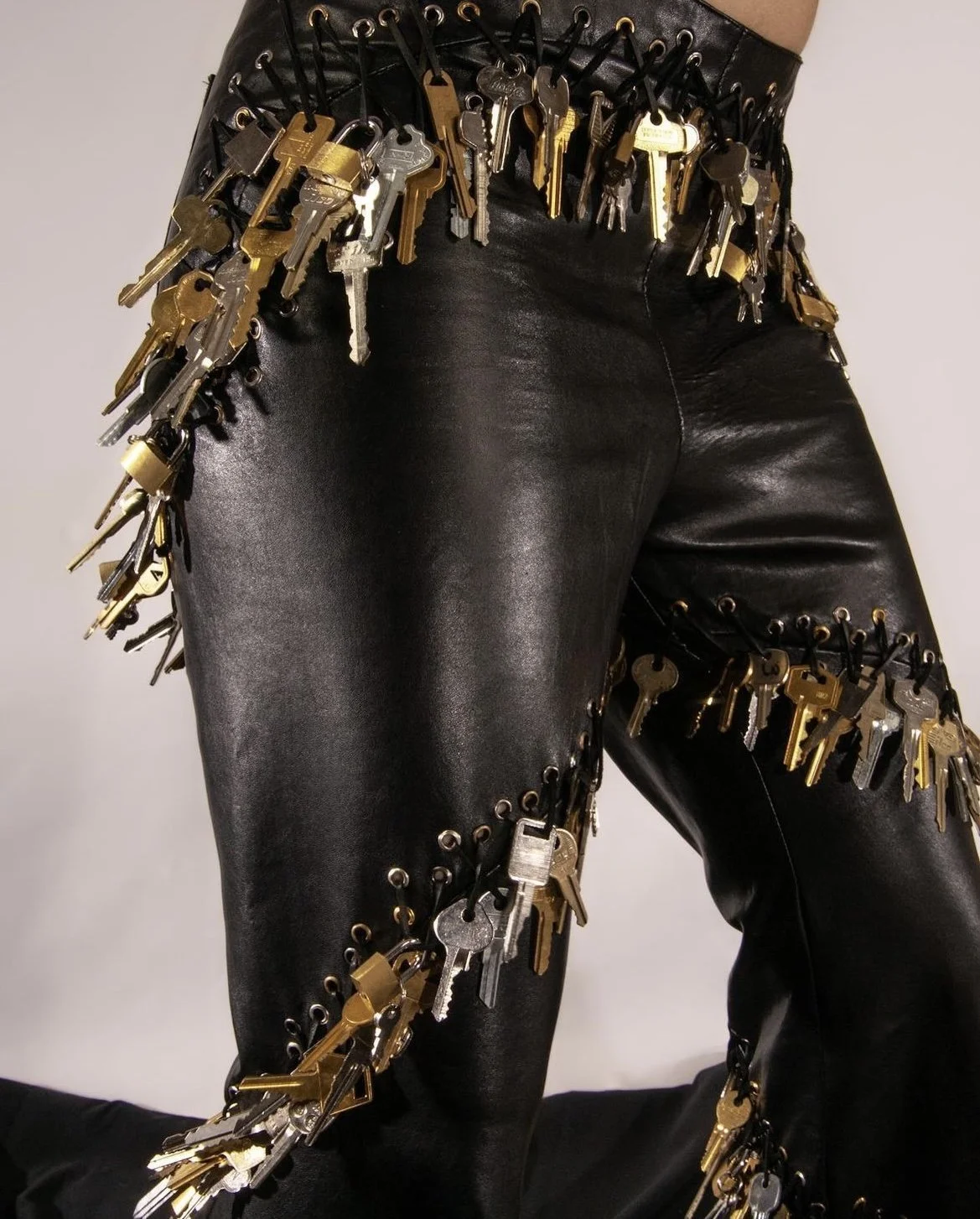 Close-up of black leather pants adorned with numerous keys hanging from the waistband, pockets, and legs, secured with black and gold hooks.