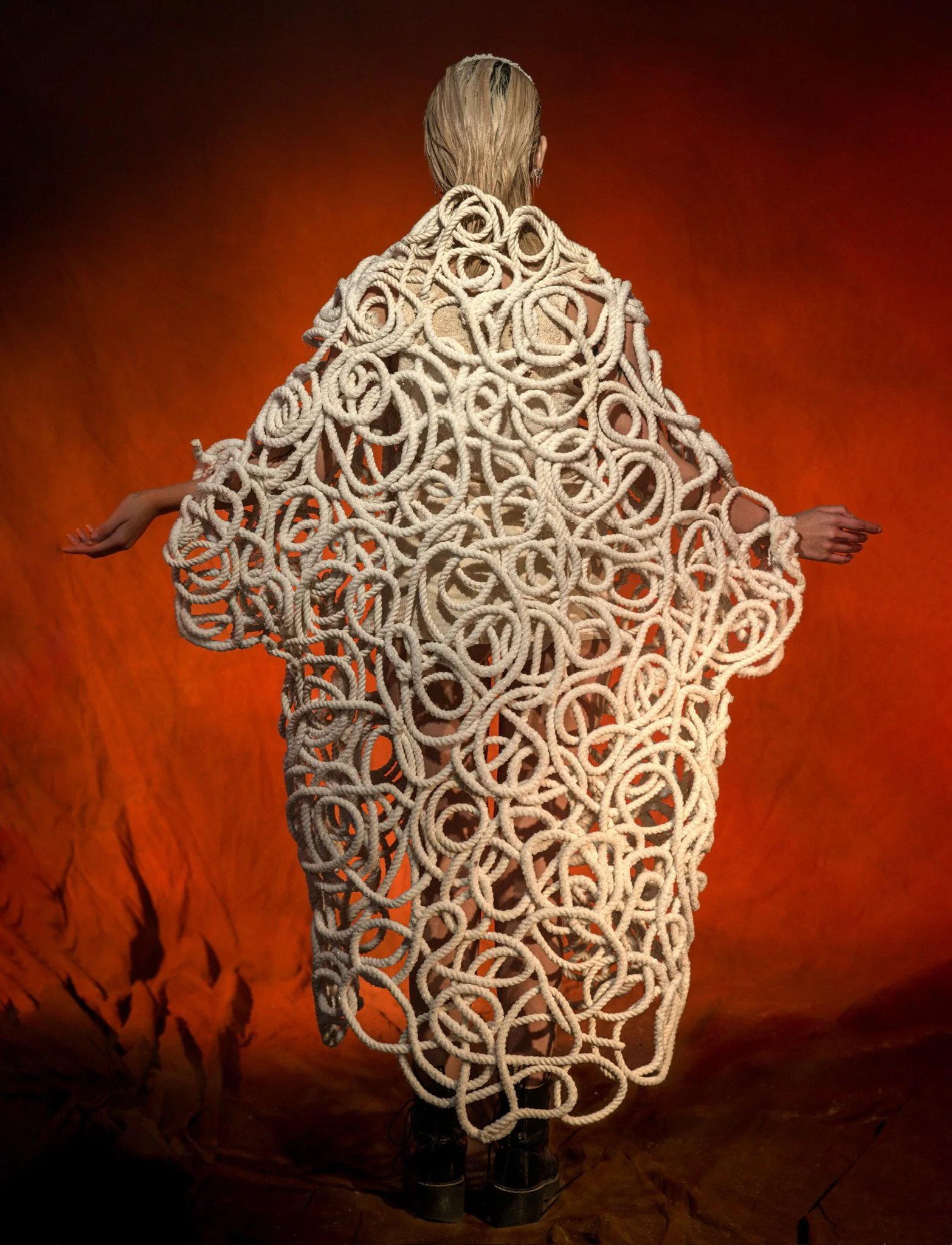 A person wearing a dramatic, oversized coat made of intertwined cream-colored ropes, standing on a dark surface against a fiery orange background.