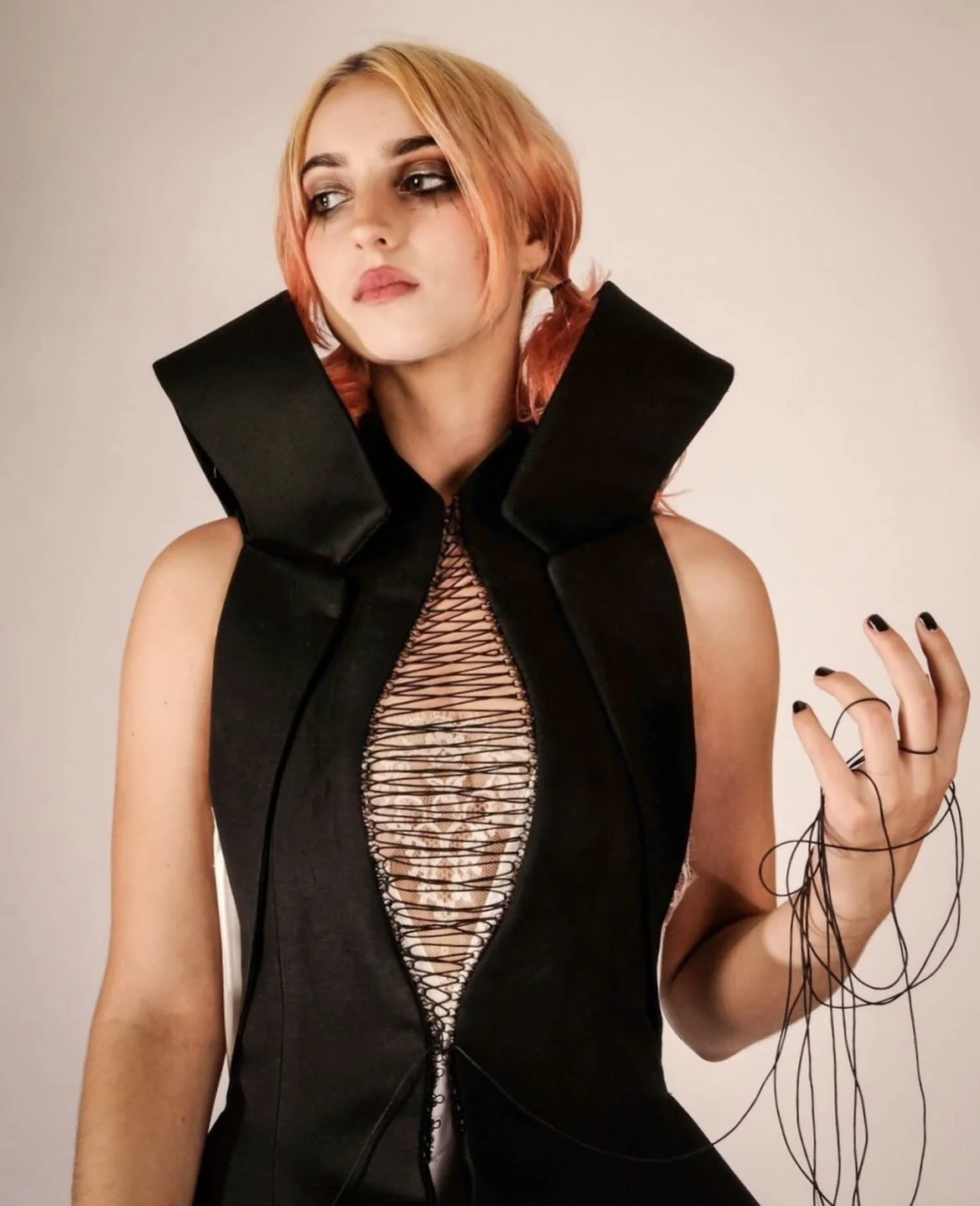 A woman with peach-colored hair wearing a black, high-collared top with cutout lace detail is holding several black wires or cords in her right hand, standing against a plain beige background.