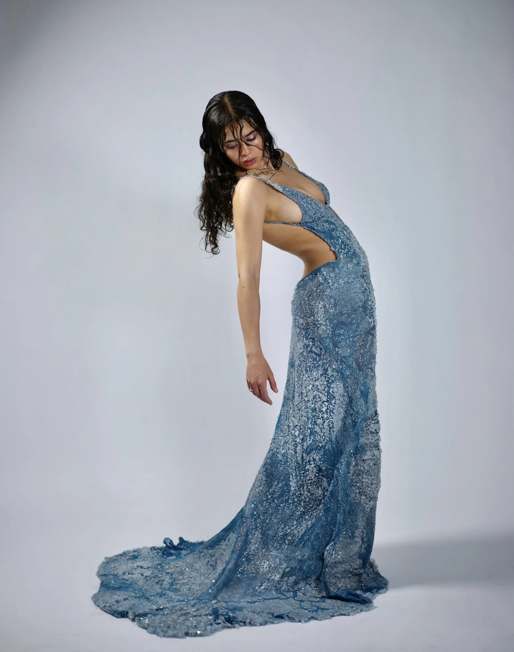 A woman with dark, curly hair is wearing a long, shimmering blue gown with cutouts on the sides, standing against a plain light background.