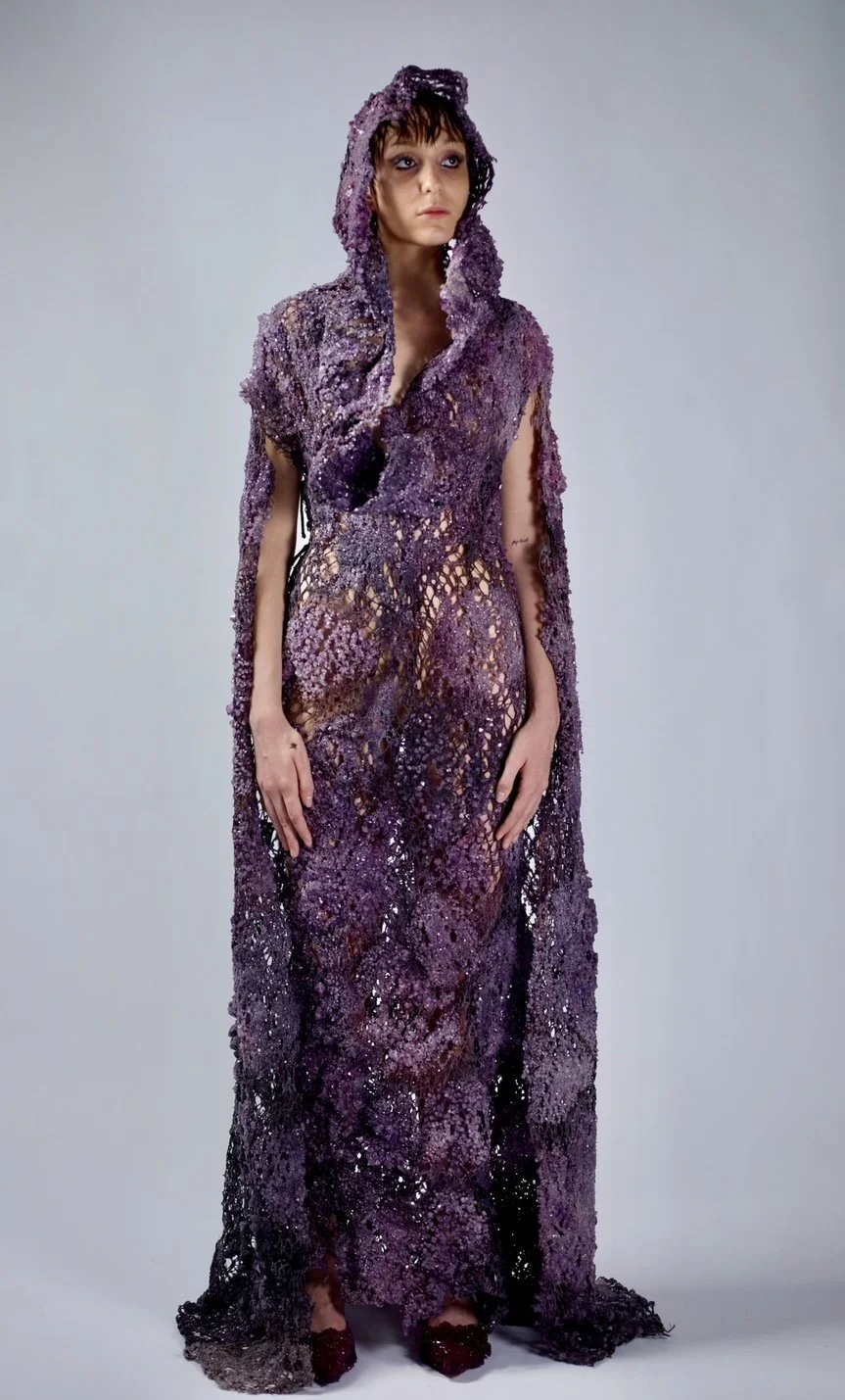 A woman wearing a purple, textured, semi-sheer gown with a hood, standing against a plain background.
