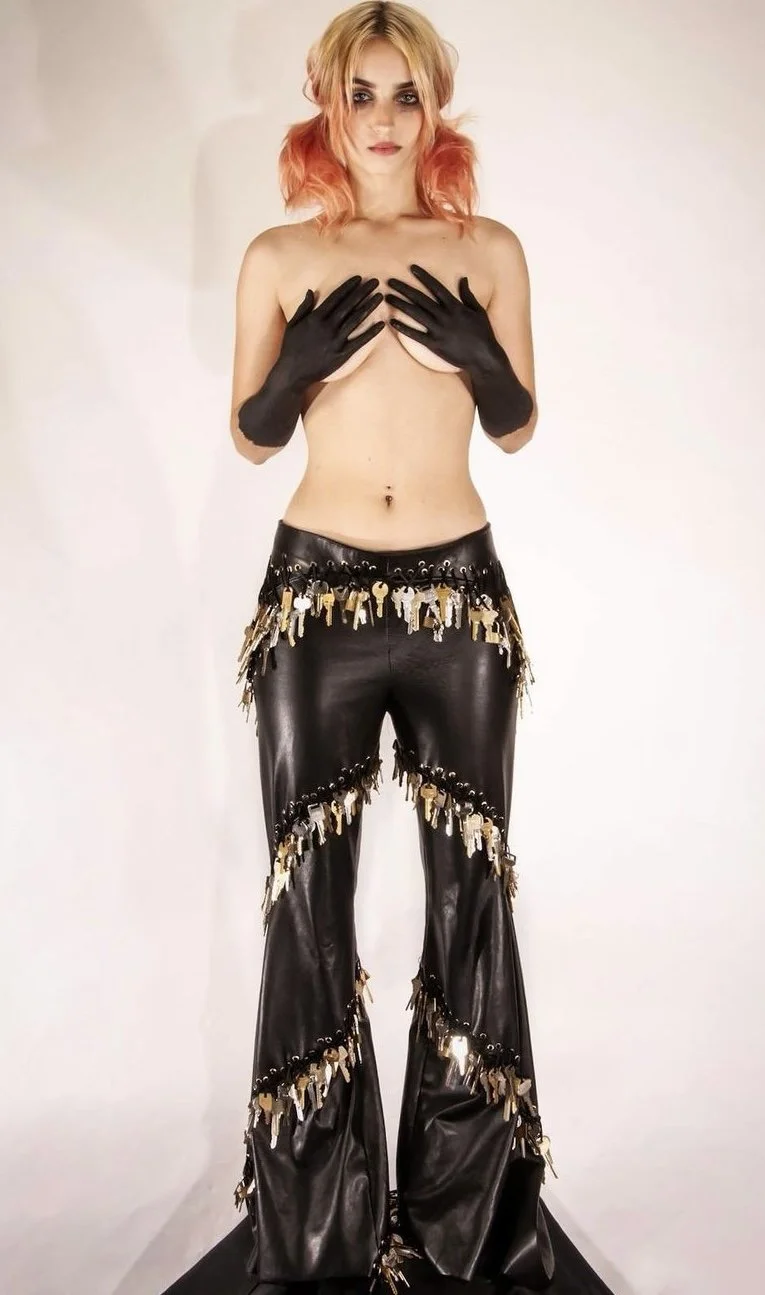 A woman with pink and blonde hair, wearing black gloves and black leather pants with fringe and keychain decorations, is topless and covering her chest with her hands.