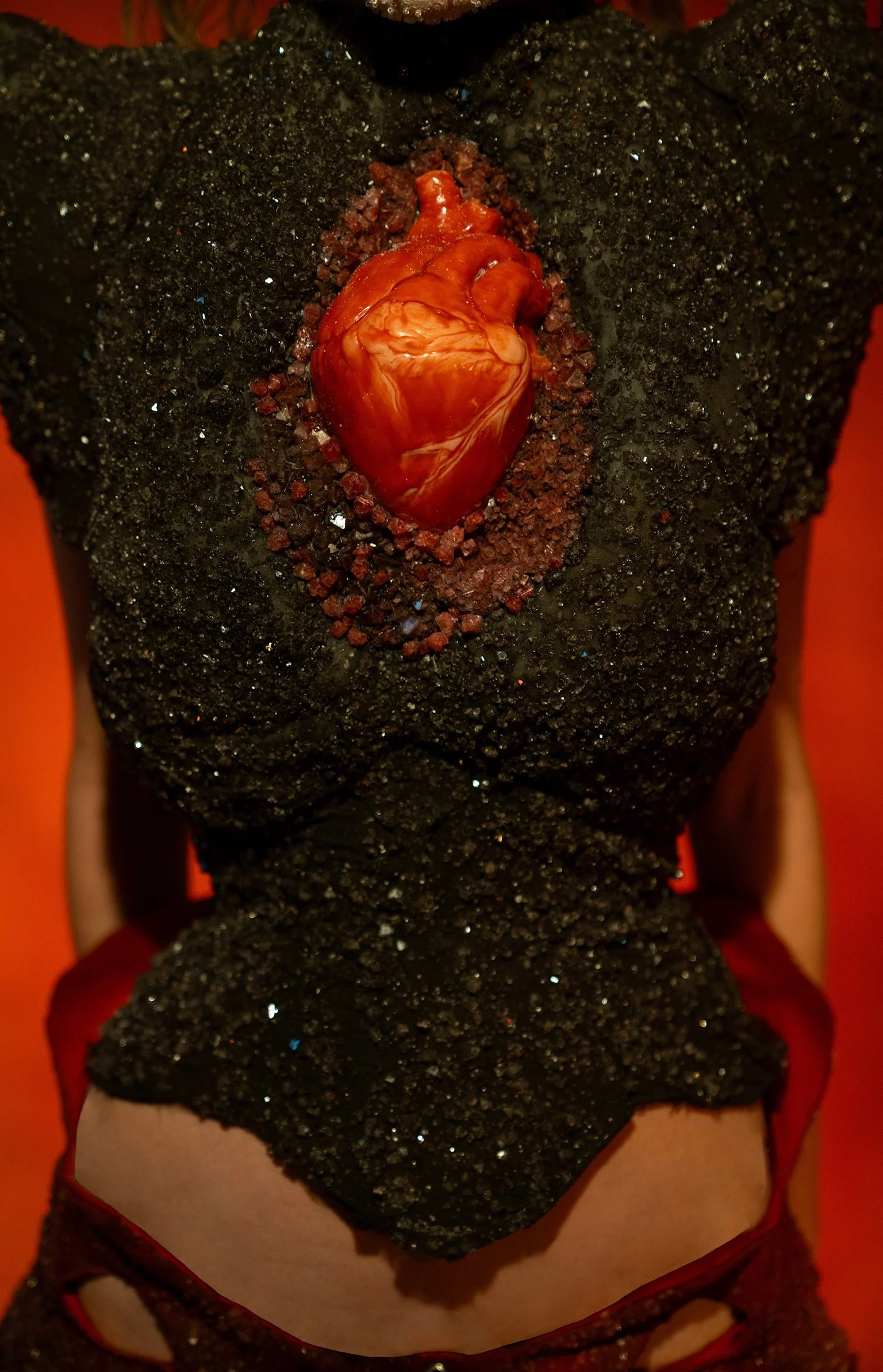 Close-up of a person wearing black glittery dress with a heart-shaped red gemstone on the chest, seen from the neck down.