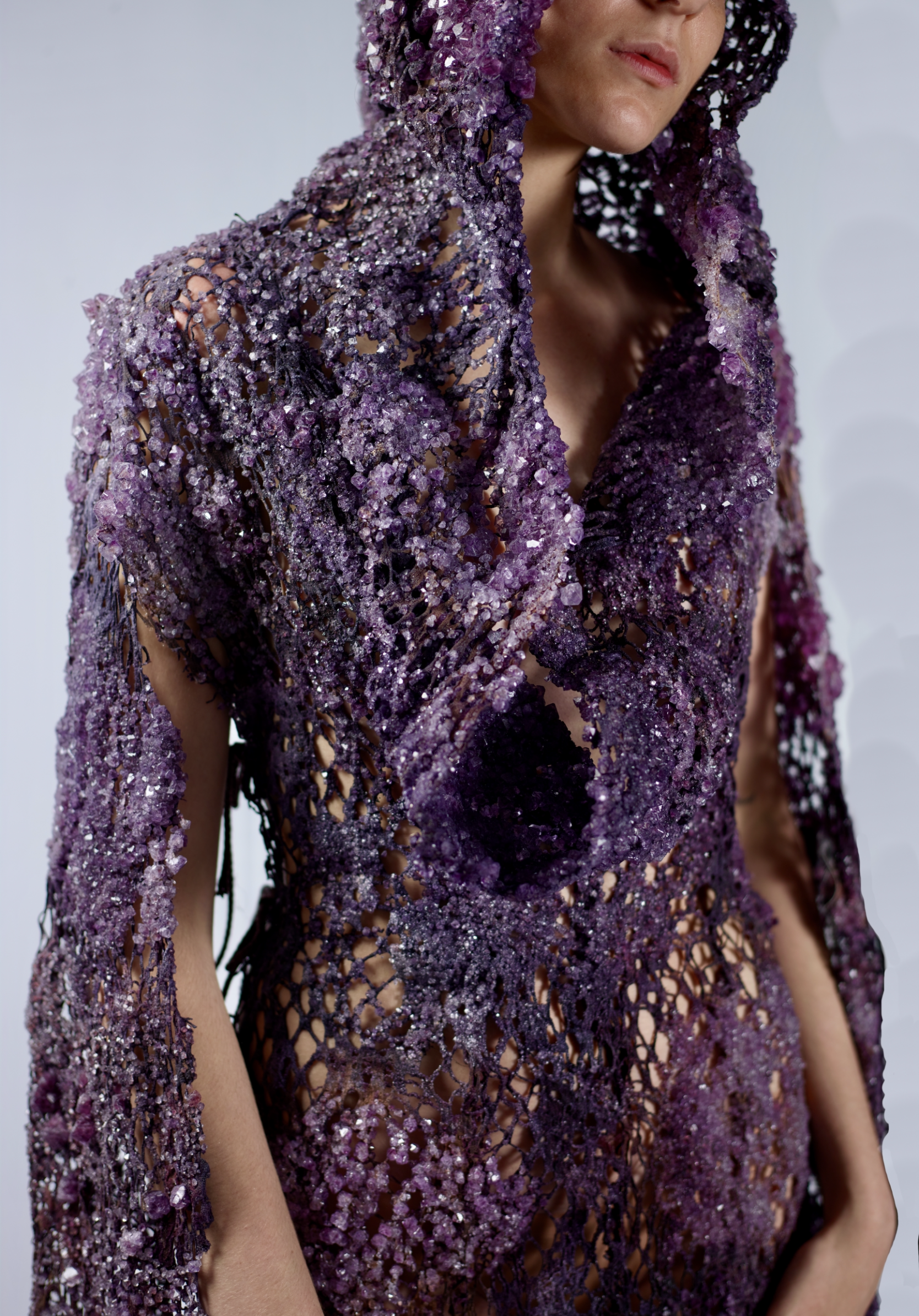 A person wearing an intricate hooded garment made of purple crystals and beads, with an open weave design revealing their skin underneath.