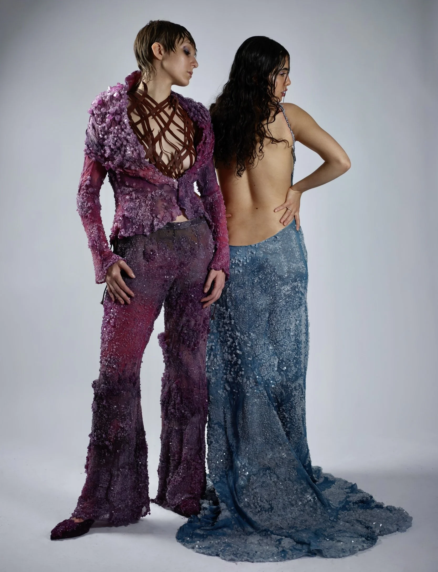Two women dressed in glittering, textured gowns with one in pink and purple hues and the other in blue, standing against a plain white background.