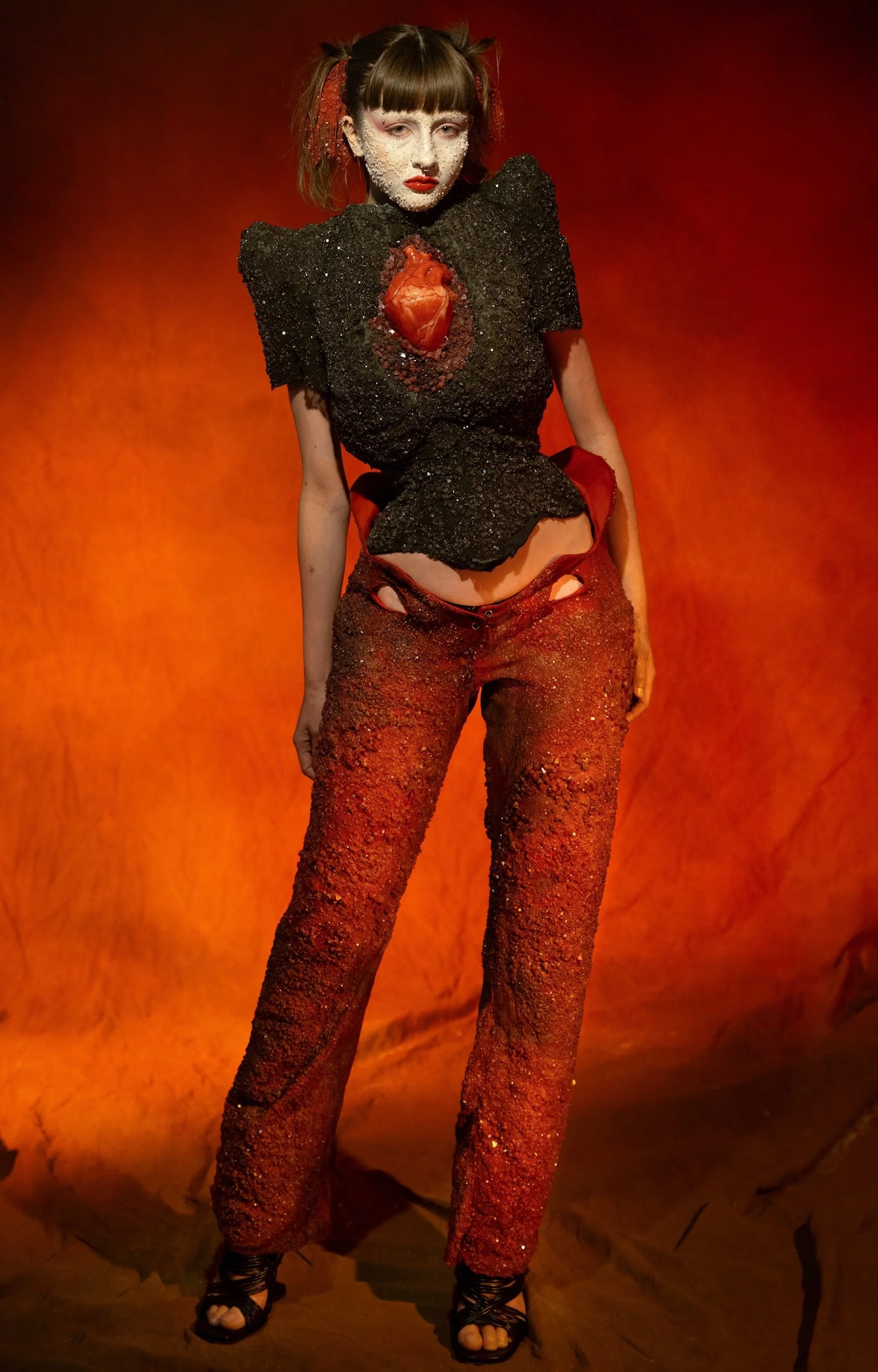 A model wearing a black textured top with large shoulder pads and a red heart in the center, paired with red textured pants, standing against an orange background.