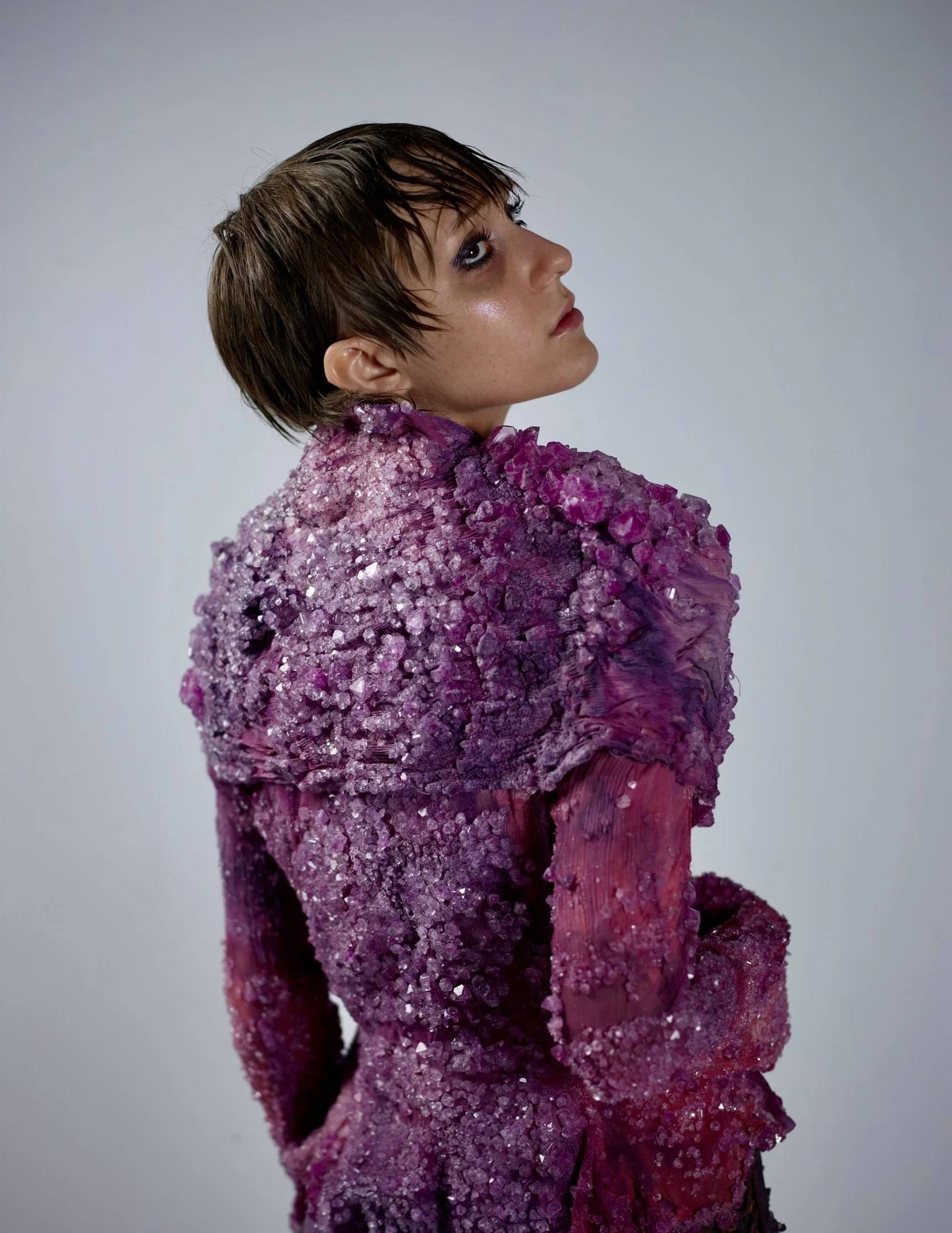 Person with short brown hair wearing a purple and pink textured dress with crystals and ruffles, looking to the side against a plain background.